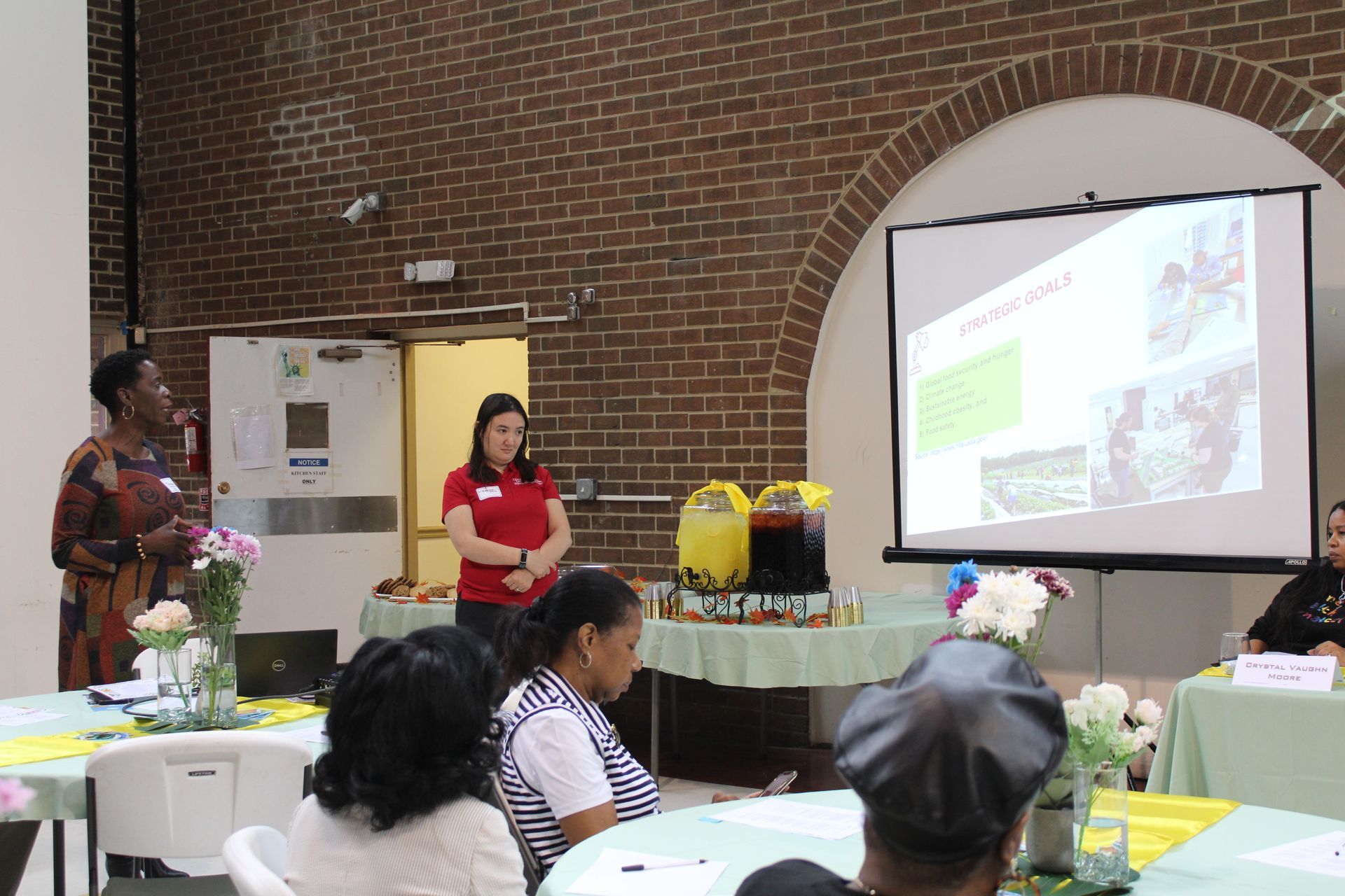Two women present to an audience; a slideshow on a screen. Tables are set with flowers.