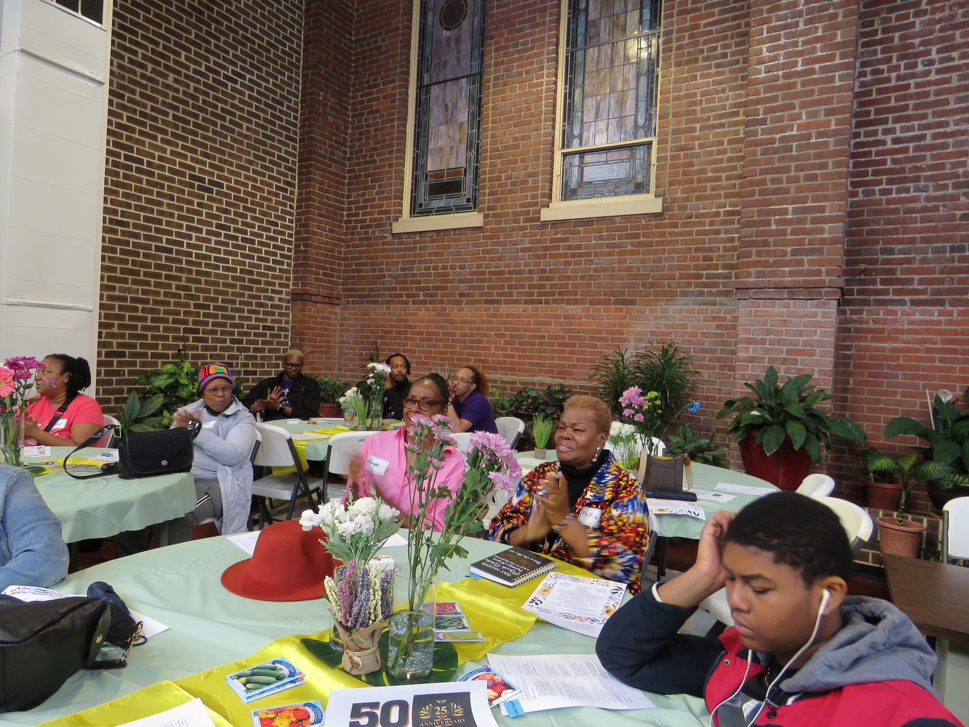 People seated at tables in a room with brick walls, some looking at notes or laptops, flowers on the tables.