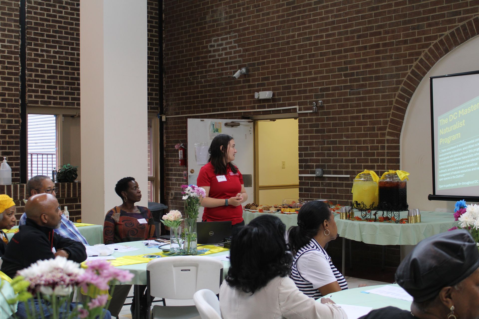 Woman in red shirt presenting to a seated audience at a meeting, projector screen in the background.