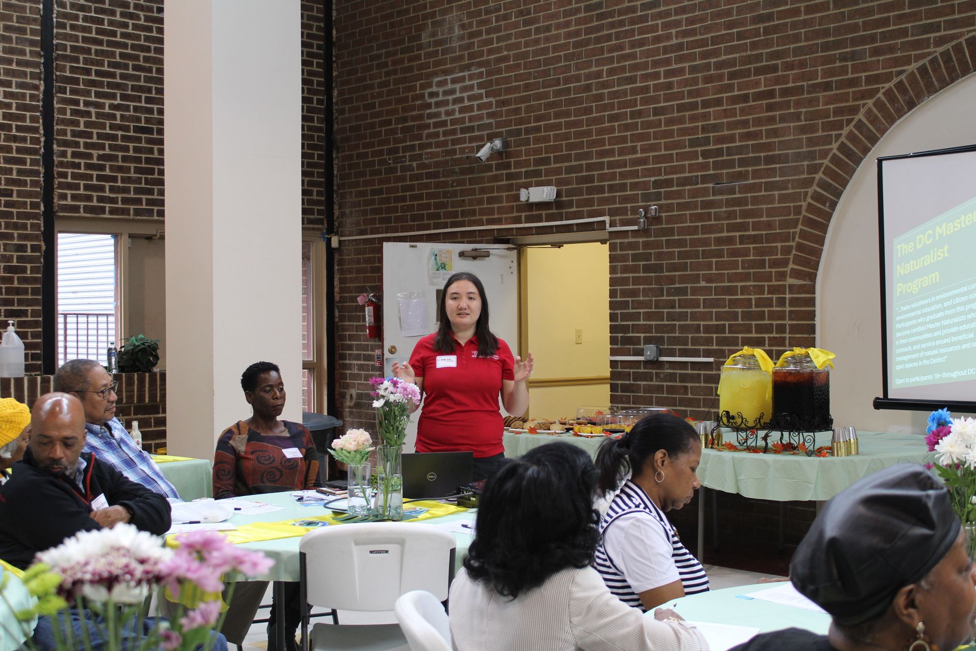 Woman in red shirt presenting to a seated audience at a community event, with food and a projector.