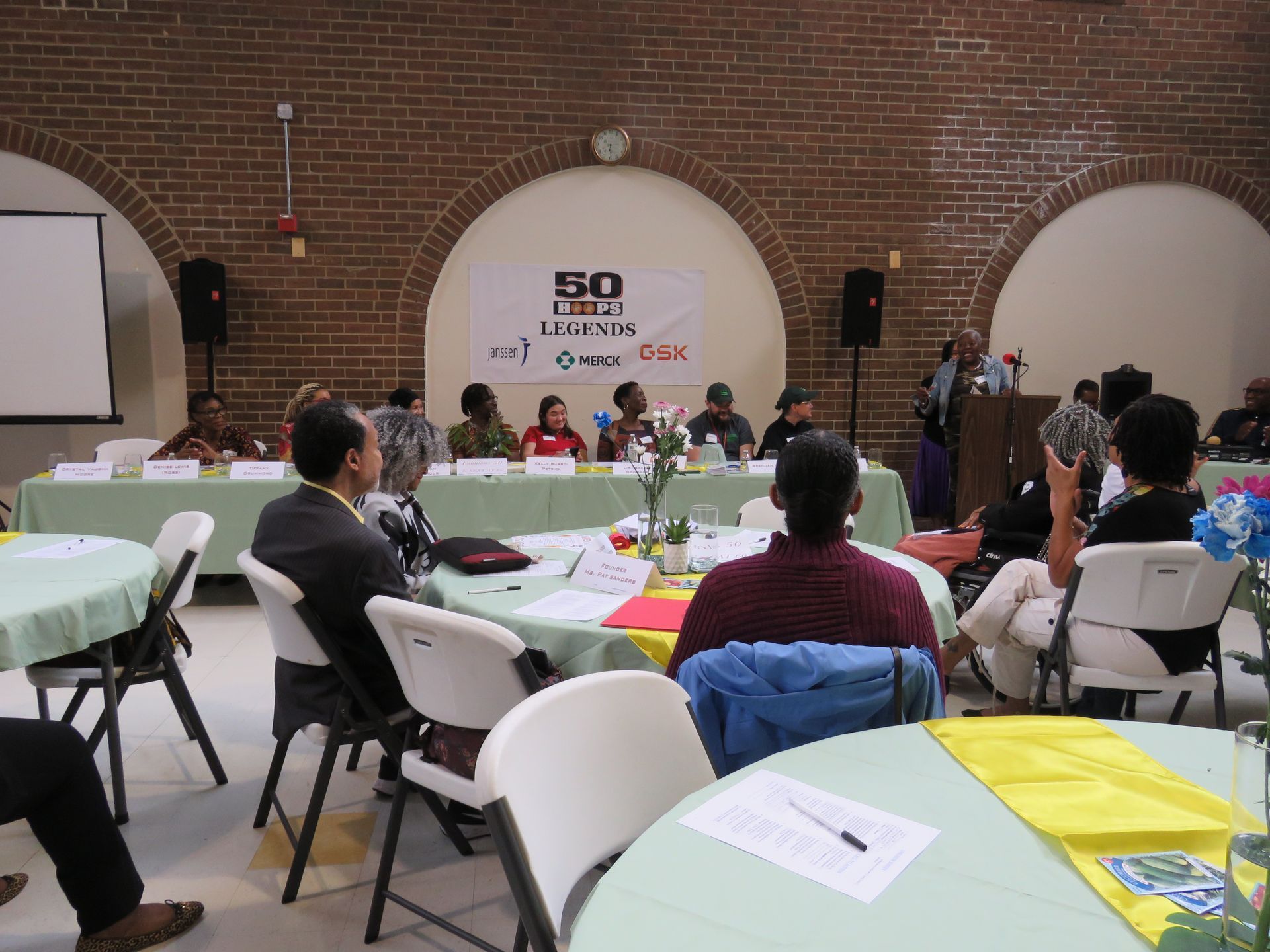Panel discussion at a conference, speakers behind a long table, audience at round tables. Brick wall background.
