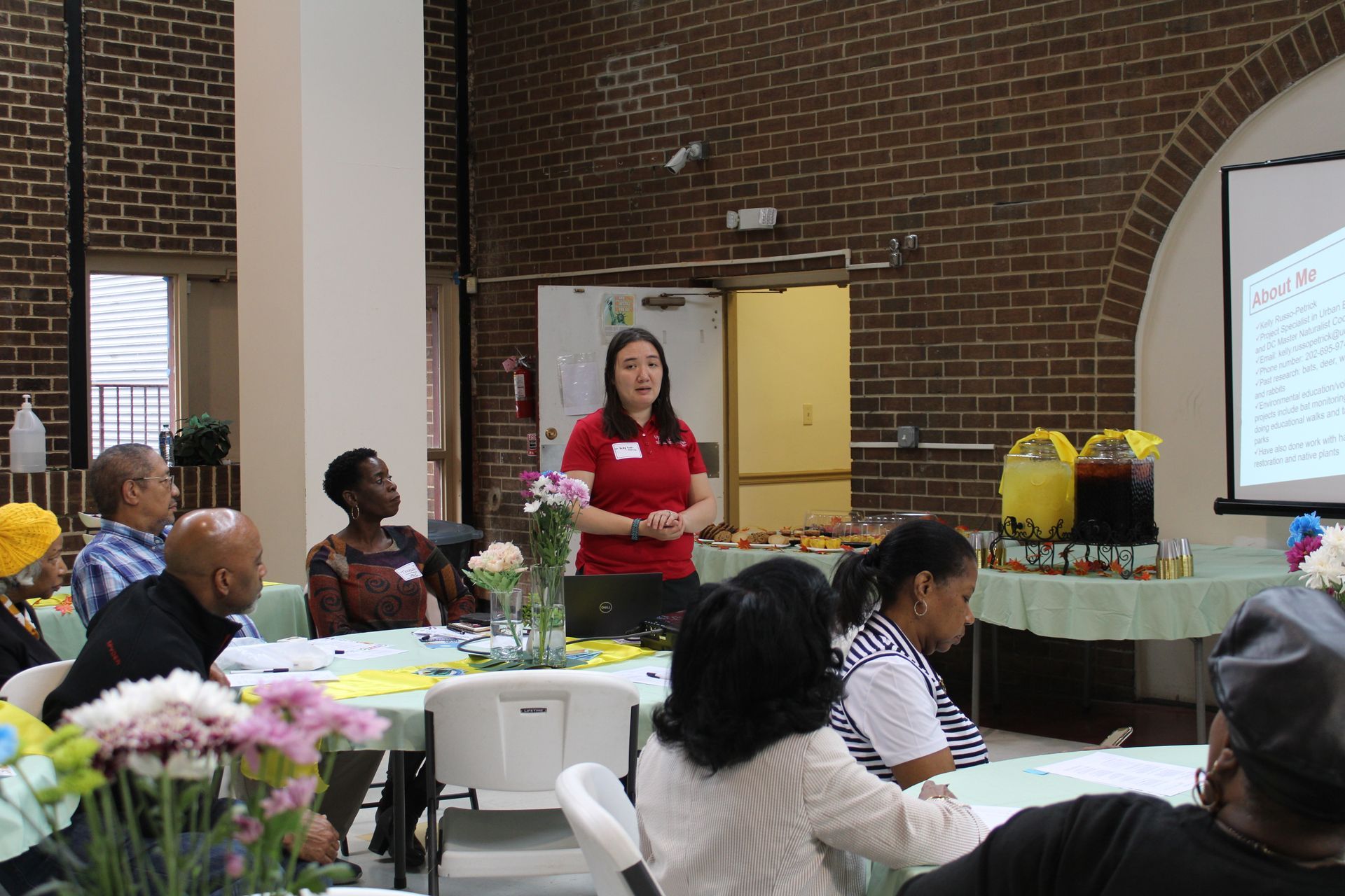 Woman presenting at a meeting, standing before a projector screen, in a brick-walled room with seated attendees.