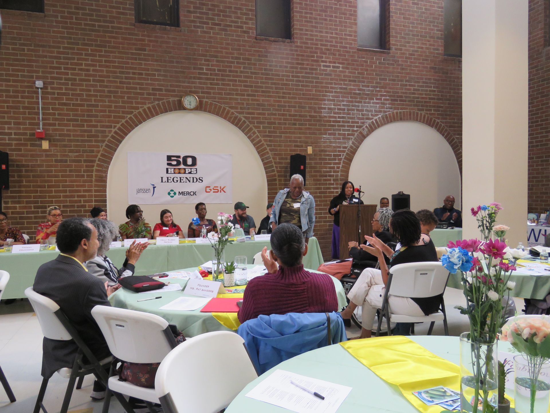 A community meeting in a brick building. People seated at tables clap while a speaker addresses the room.