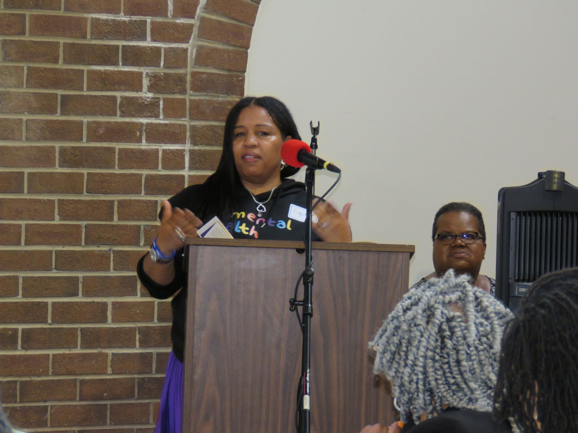 Woman speaking at a podium with microphone. Brick wall background, another person seated, audience present.