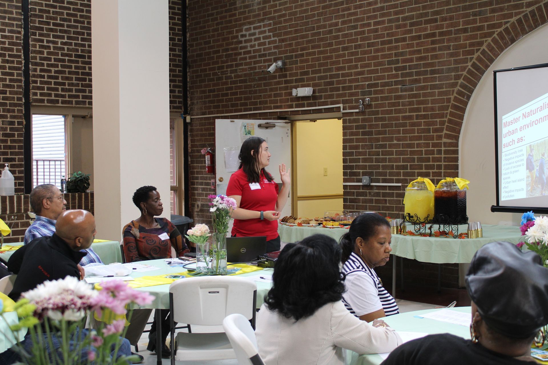 Woman presenting to seated audience at table; presentation screen visible, snacks and drinks.