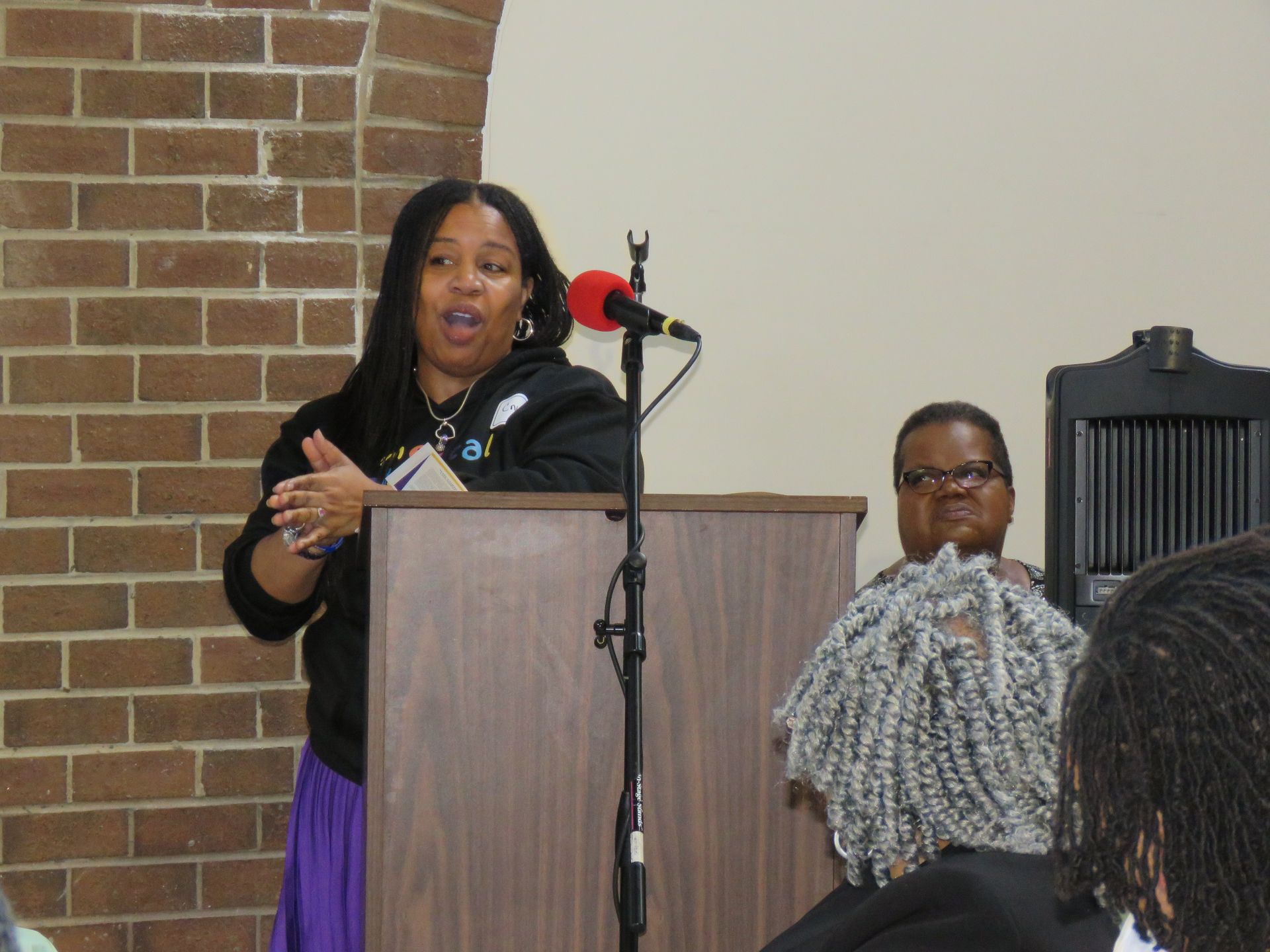 Woman speaking at a podium with microphone, gesturing with hands. Another person sits behind. Brick wall backdrop.
