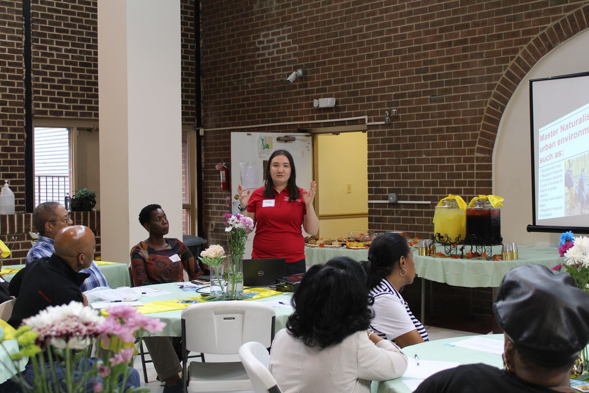 Woman presenting at a table, with people listening; a projector screen and brick wall background.