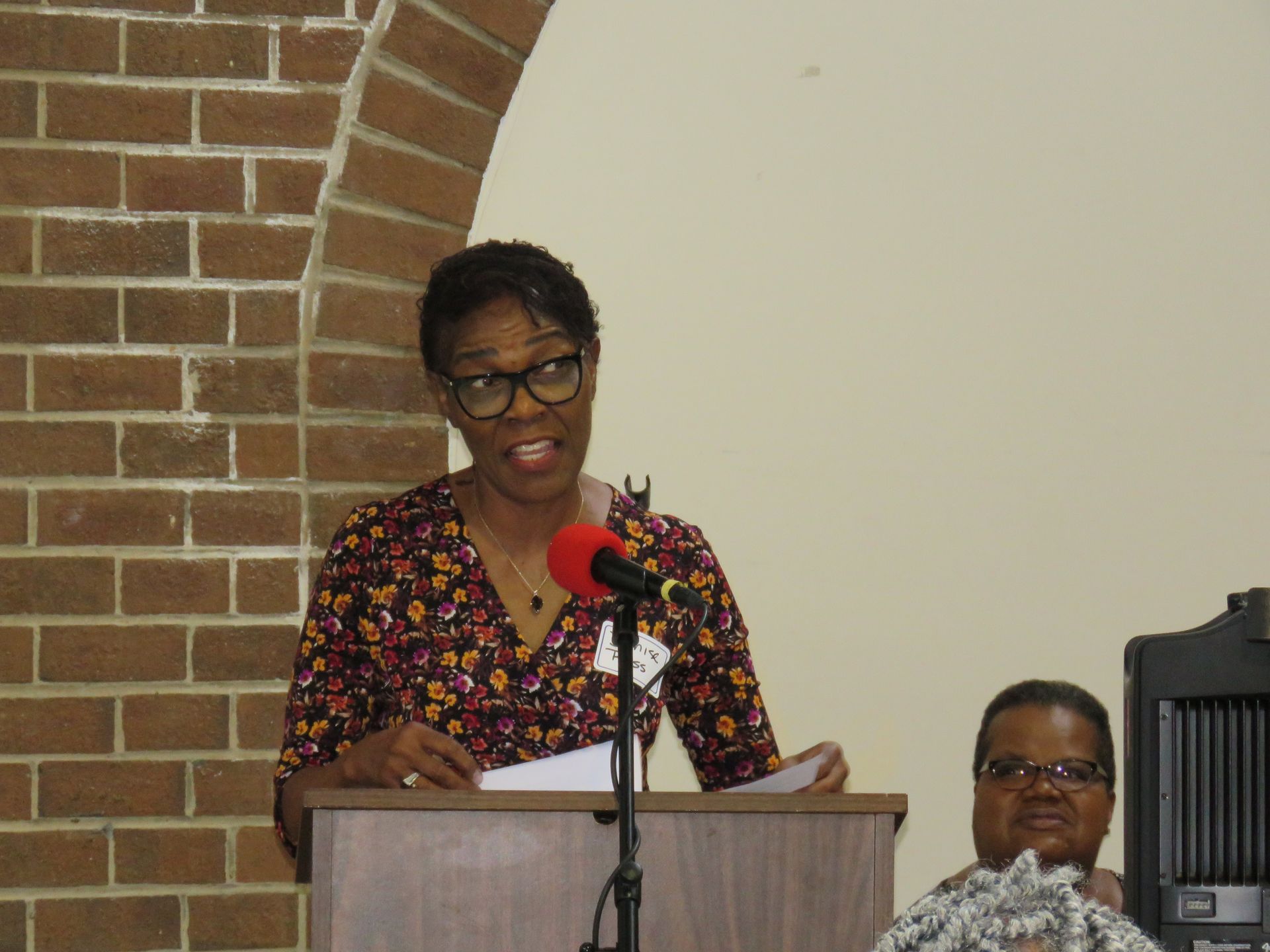 Woman speaking at a podium with microphone, papers in hand; another person seated beside her. Brick wall in the background.