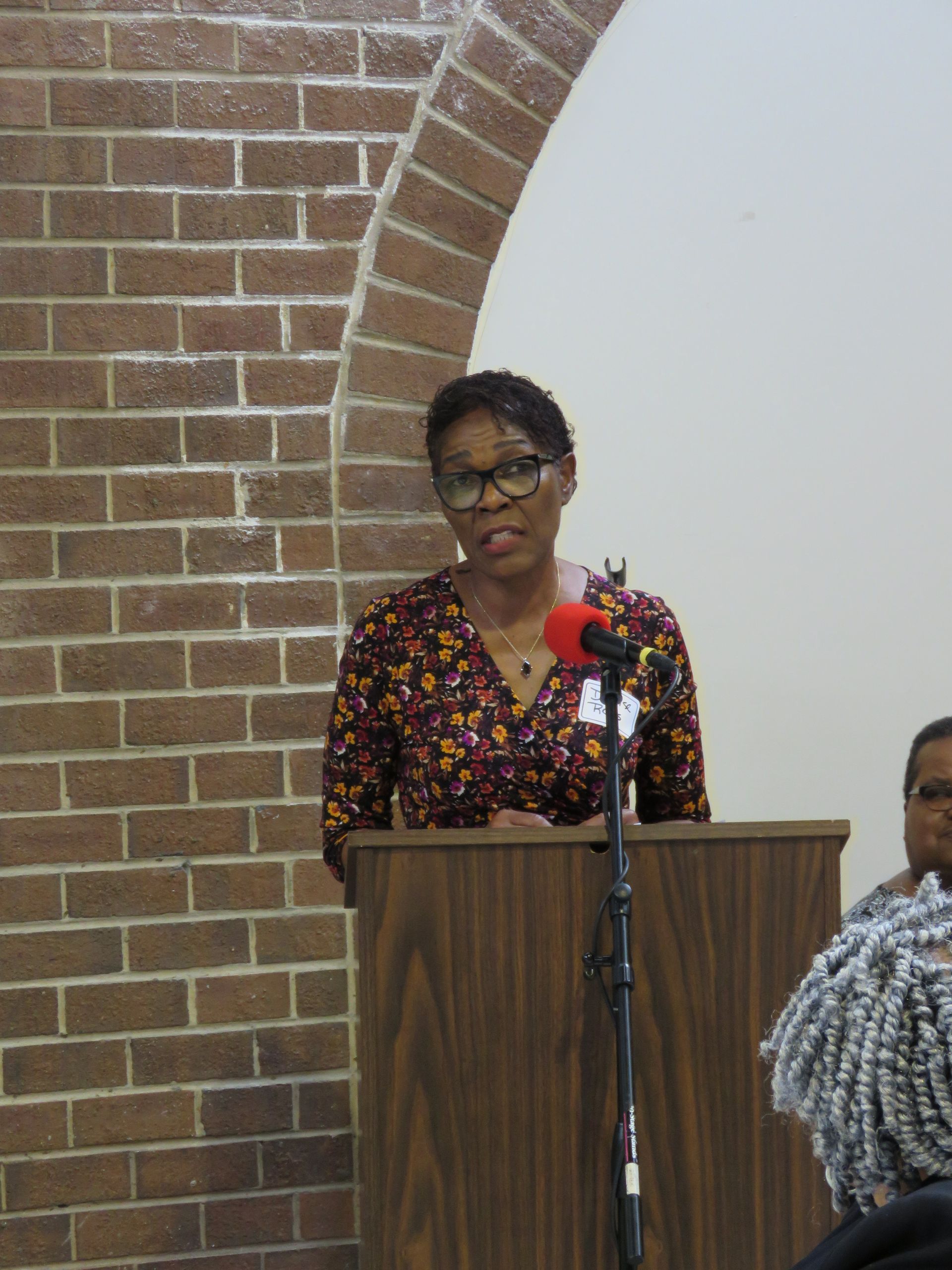 Woman speaking at a podium, near a brick archway. She is wearing glasses and a patterned blouse.