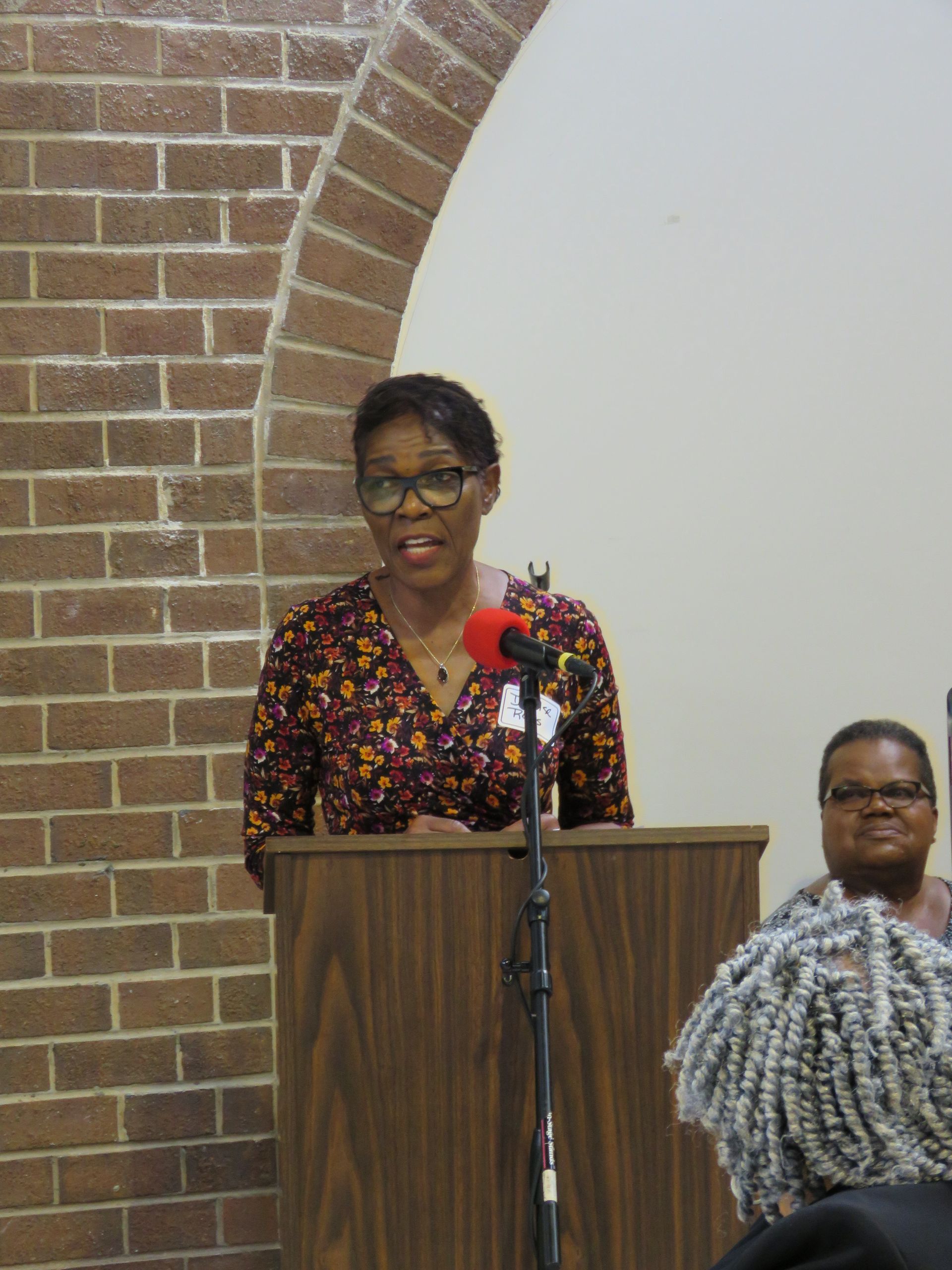 Woman speaking at a podium, brick wall and archway in the background. Another person is seated to the right.