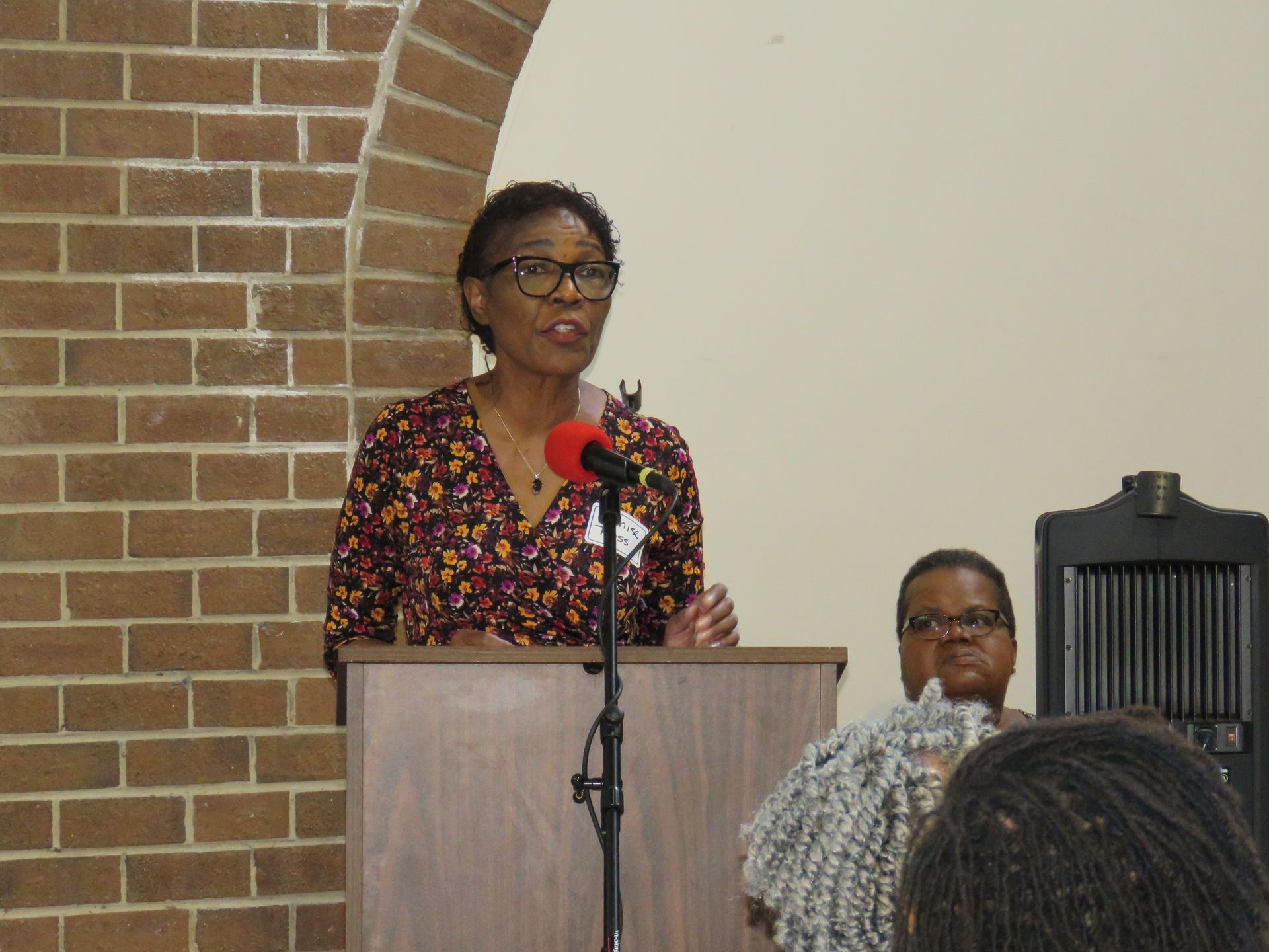 Woman speaking at a lectern, a brick wall to the left, and a person in the background.