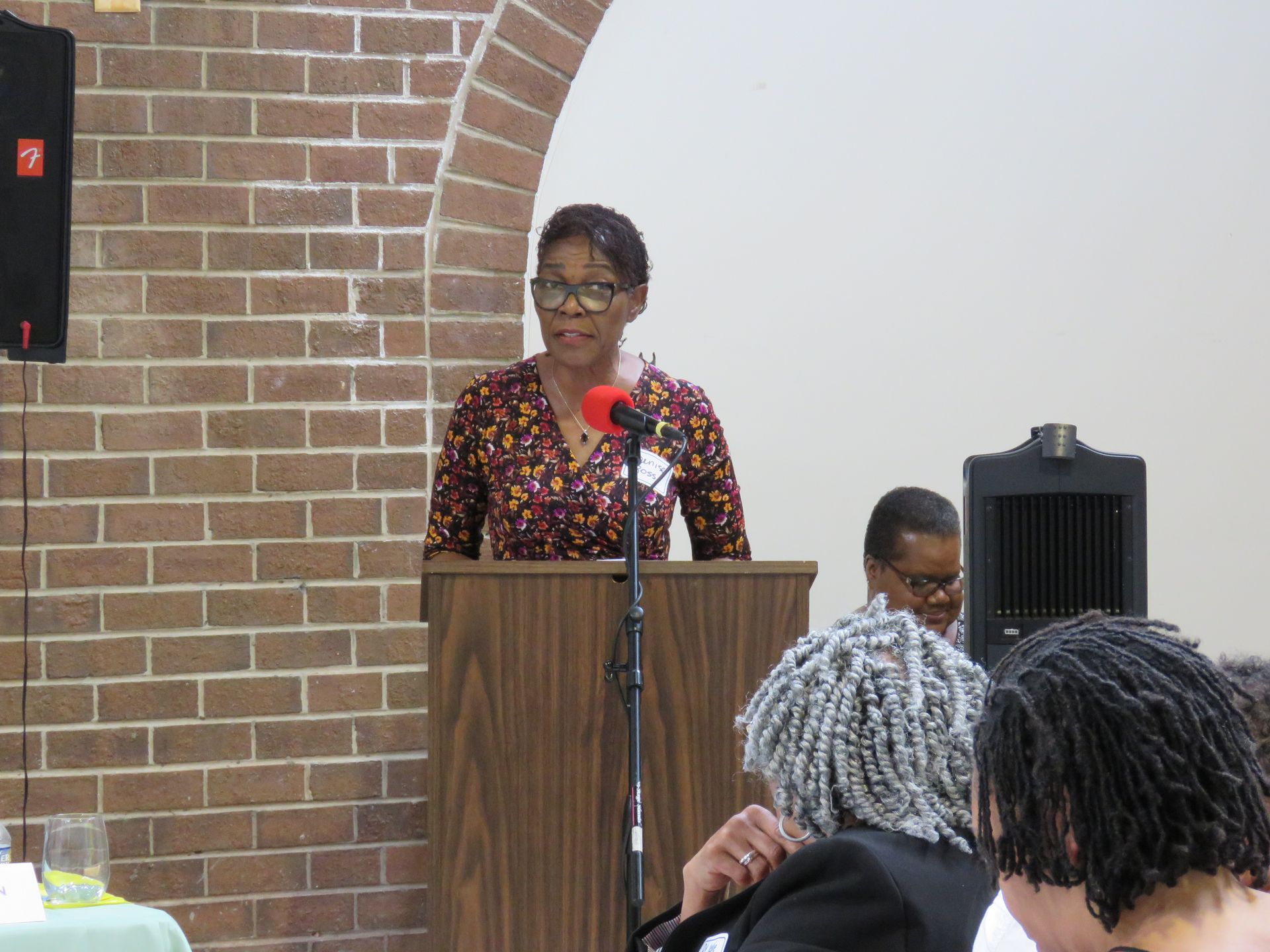 Woman speaking at a podium in front of an audience. Brick wall on the left.