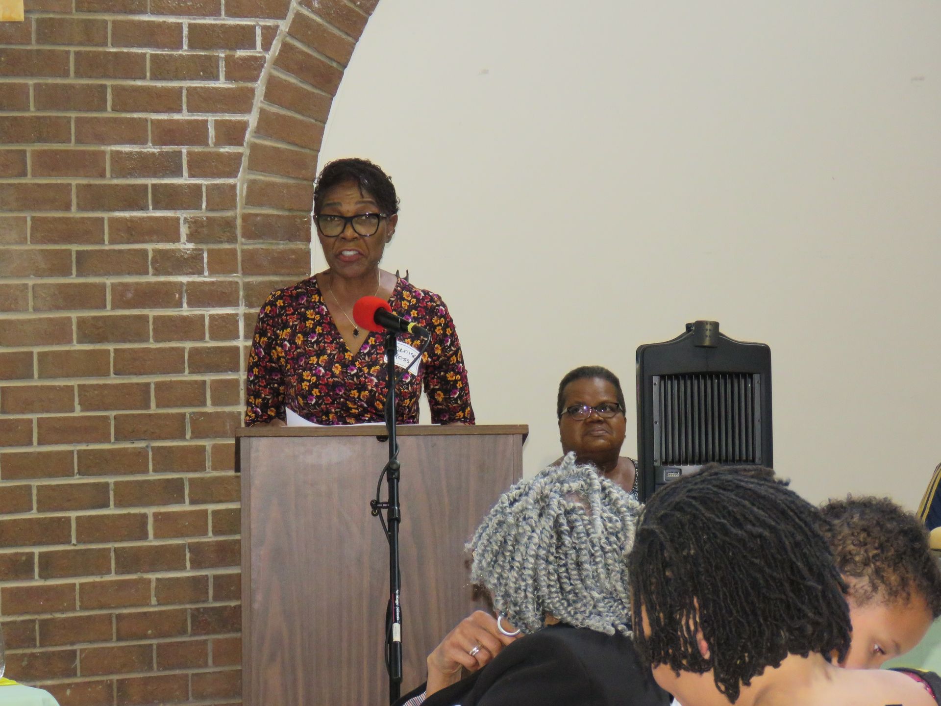 Woman speaking at a podium in a room, with people seated in the audience.