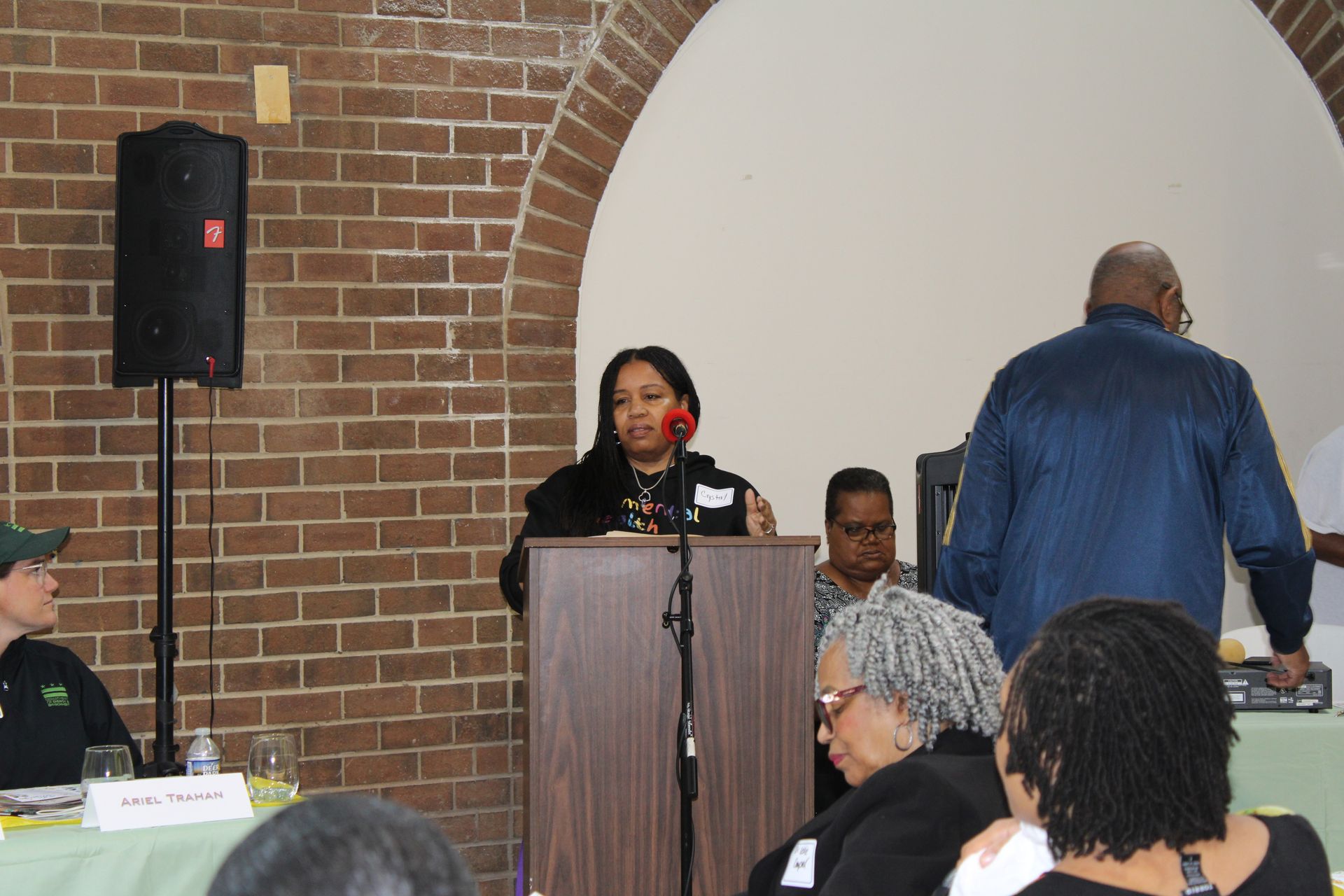 Woman speaking at a podium in a room with a brick wall and arched doorway. Others listen.