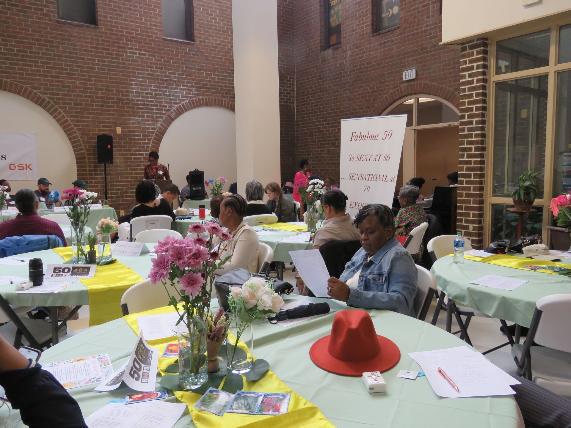People seated at round tables with flowers, attending an event in a brick-walled hall.