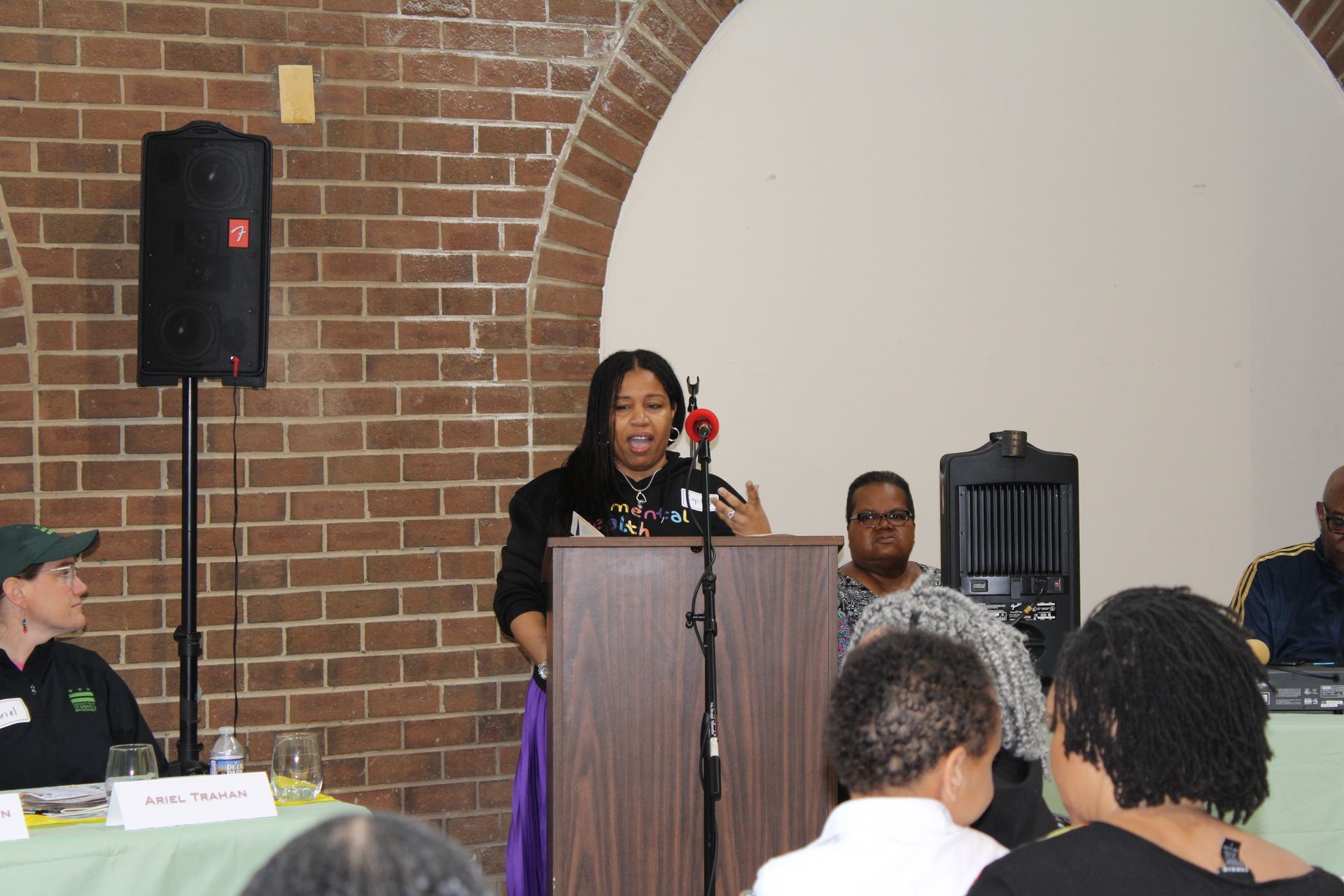 Woman speaking at a podium, brick wall backdrop. Several people seated behind a table listening.
