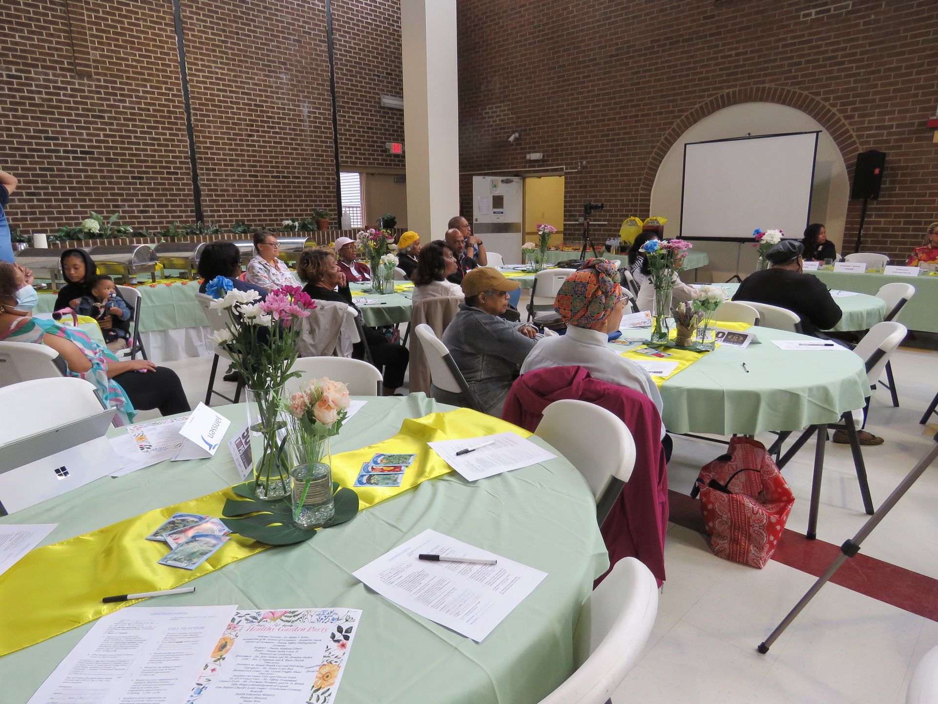 People seated at round tables in a decorated event space with a projection screen.
