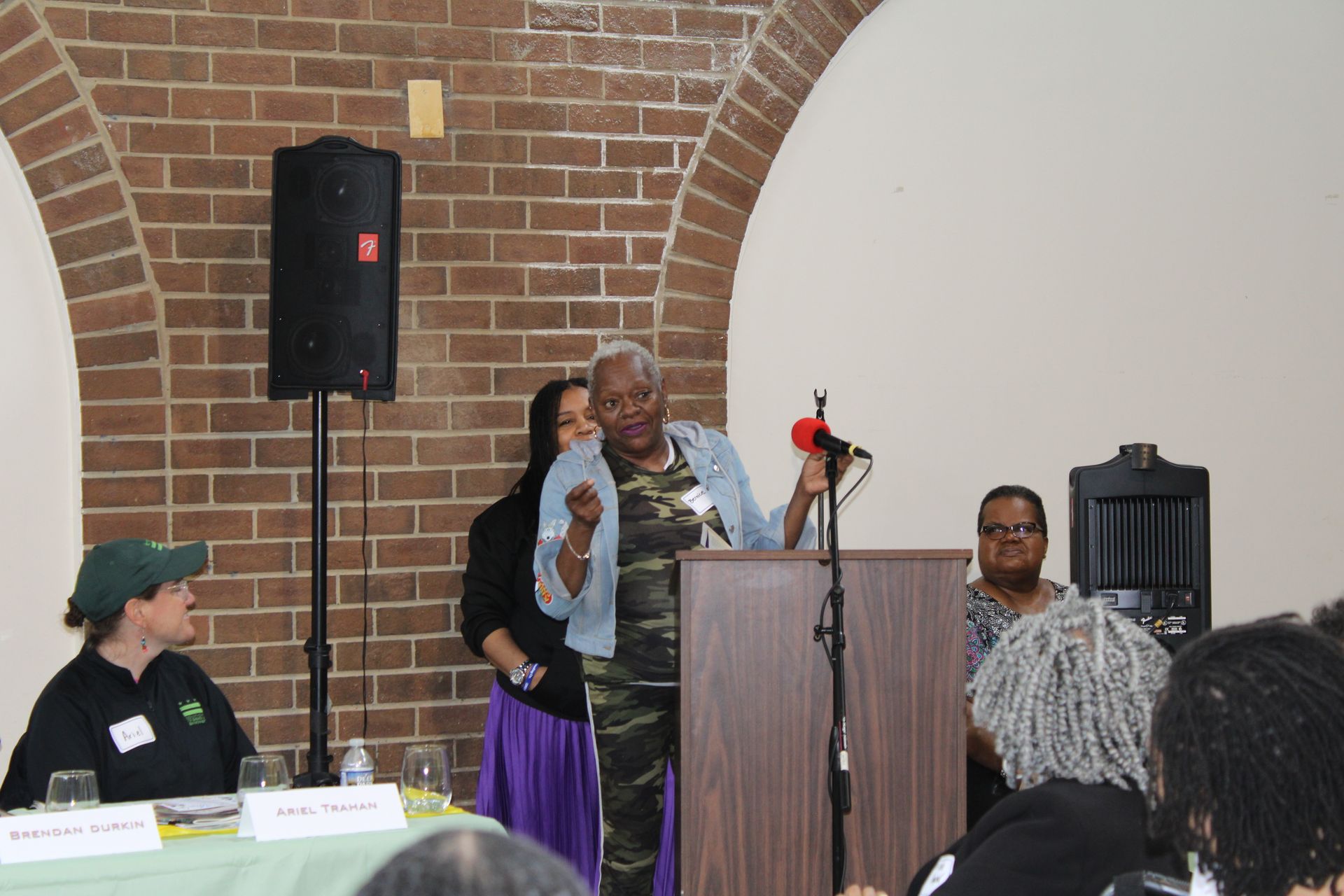 Woman speaking at a podium, others seated at a table. Brick wall background.