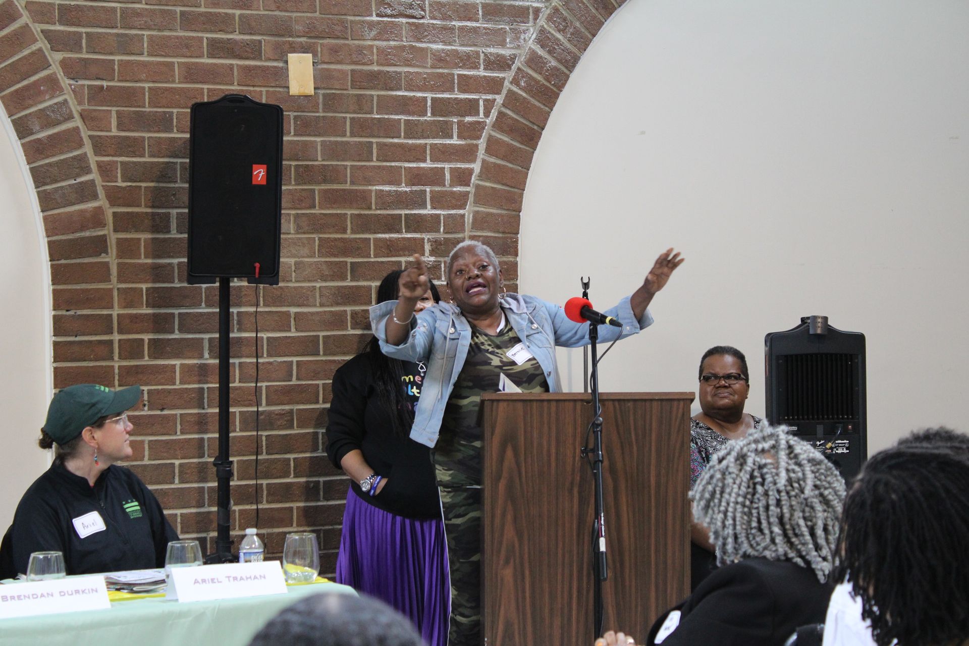 Woman speaking passionately at a podium, gesturing with arms raised. Brick wall and two other people visible.