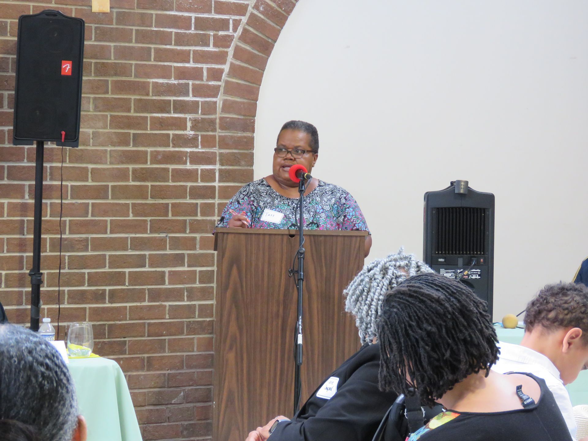 Woman speaking at a podium in front of a brick wall, with audience members seated.