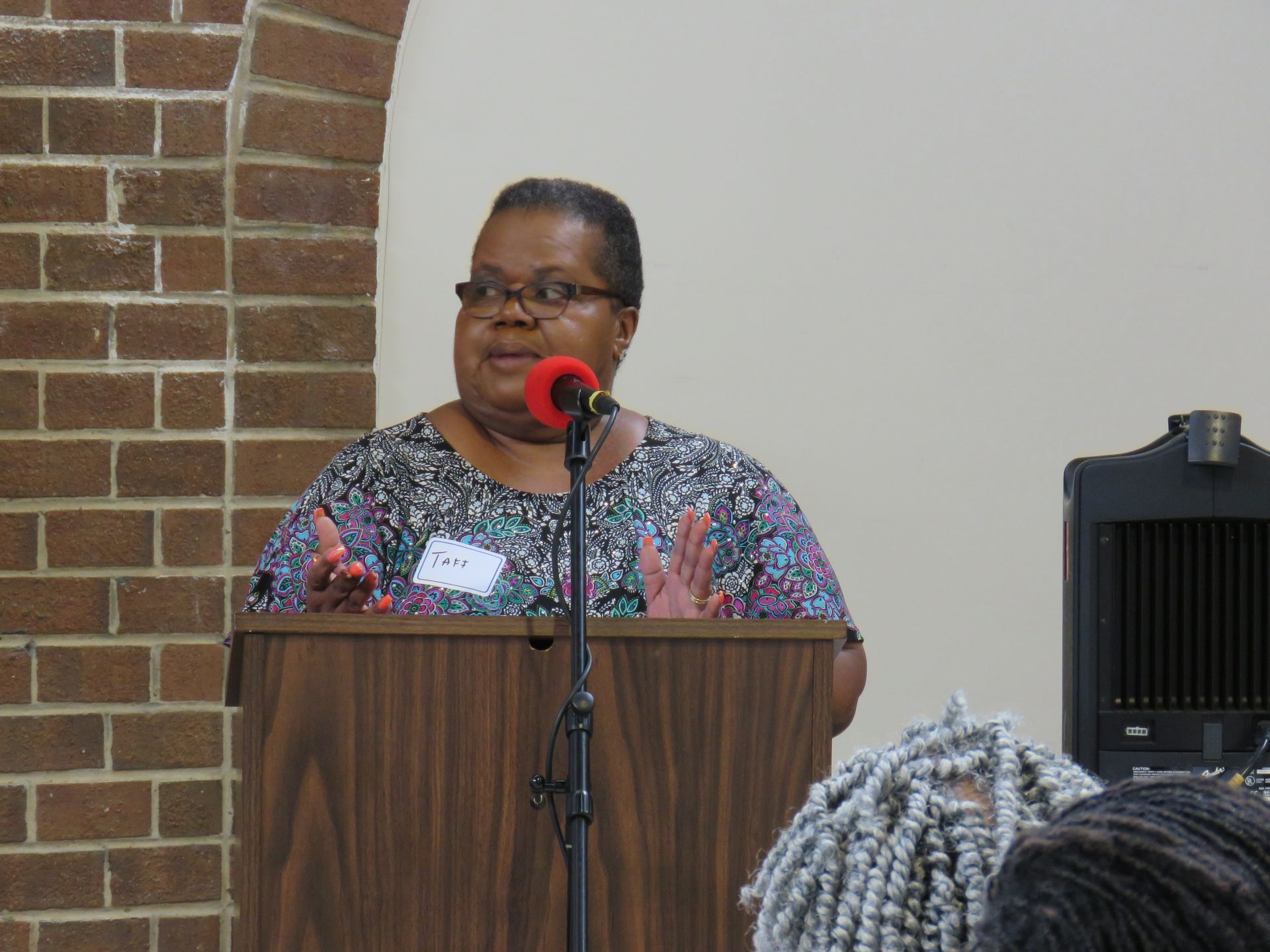 A woman speaking at a podium, gesturing with hands. Brick wall and speaker system visible.