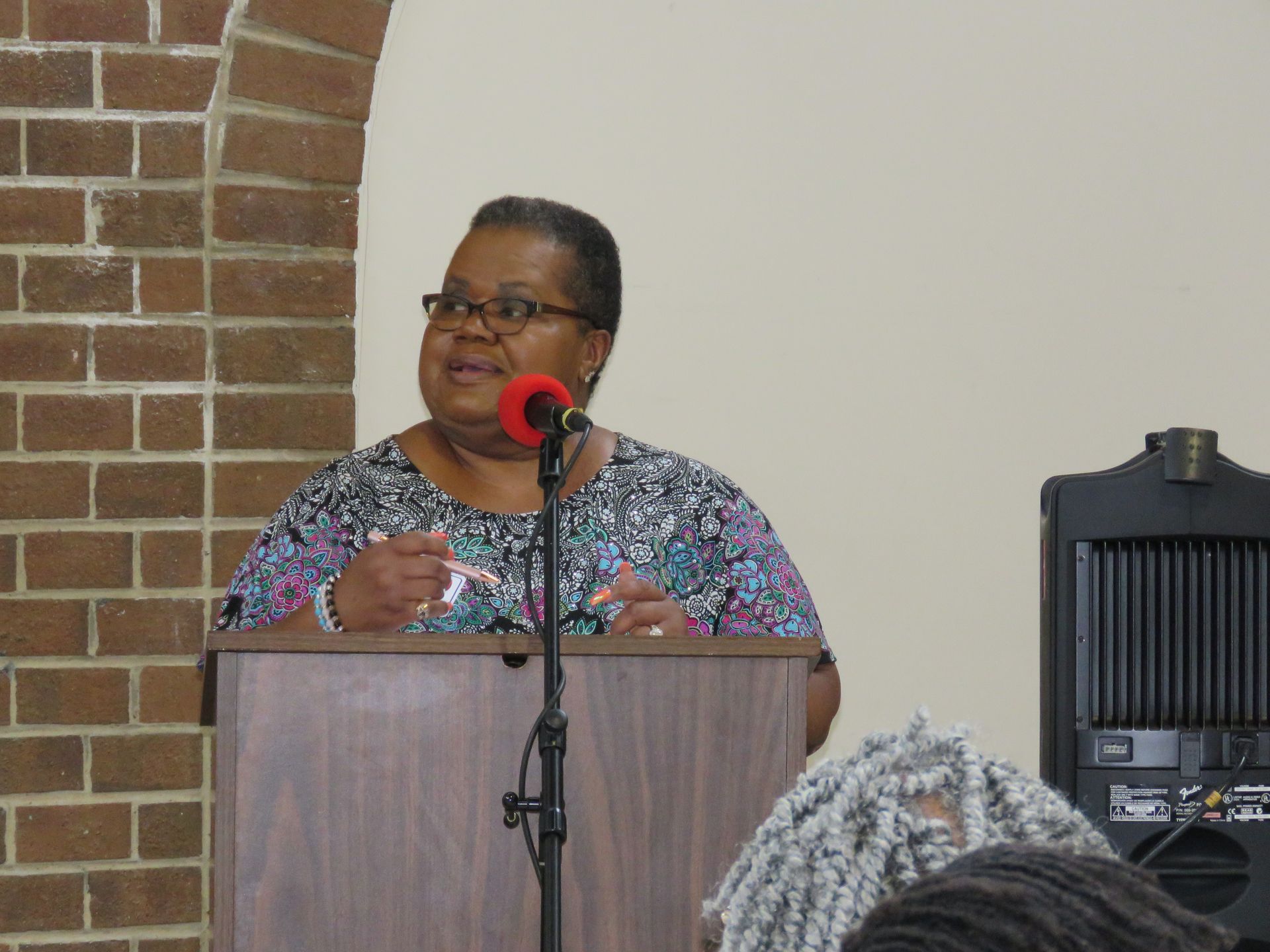 Woman speaking at a podium, wearing glasses and a patterned top, in front of brick and white walls.
