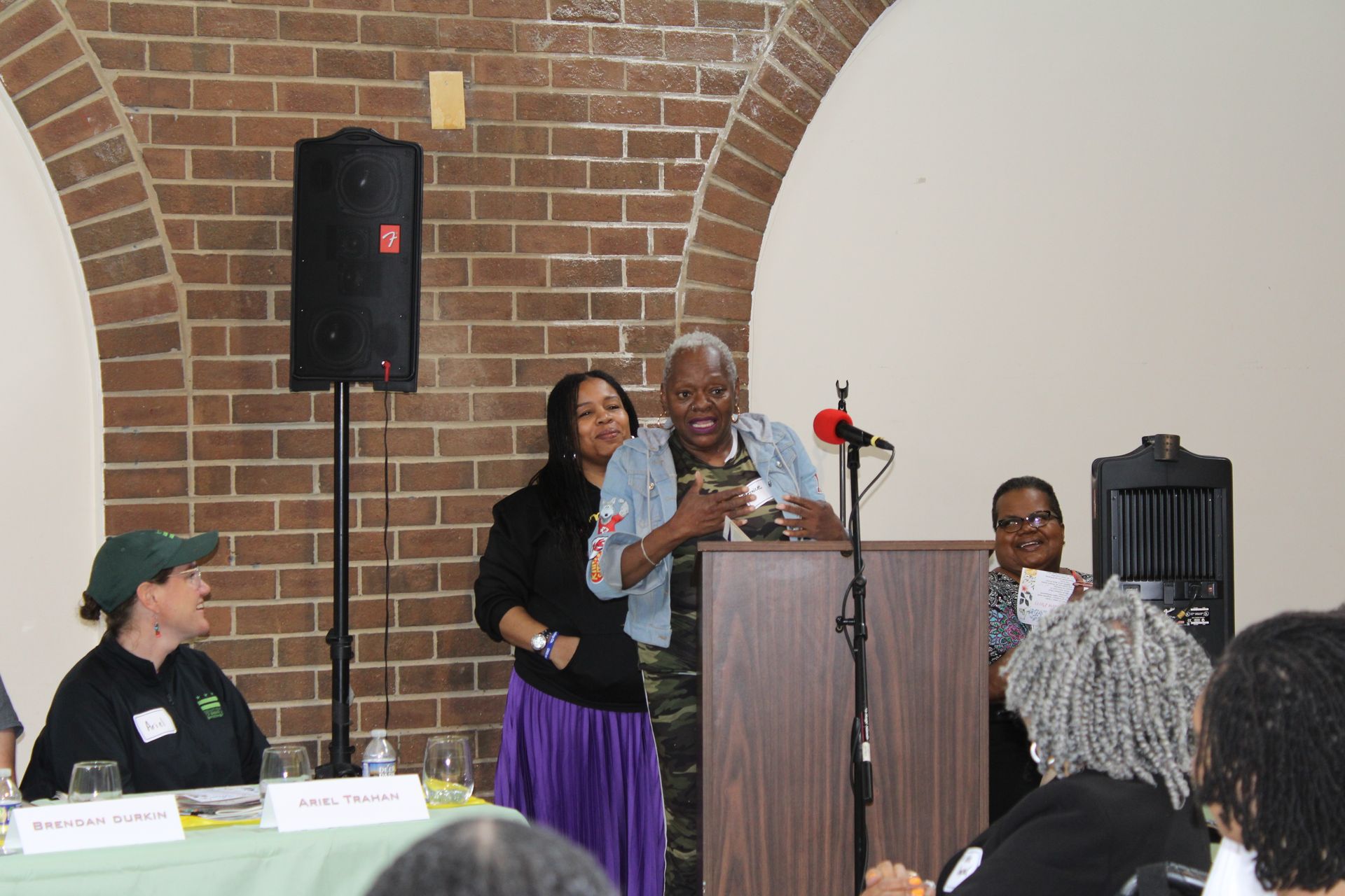 Woman at podium with microphone, holding an item, flanked by two others. Brick wall and other attendees in background.