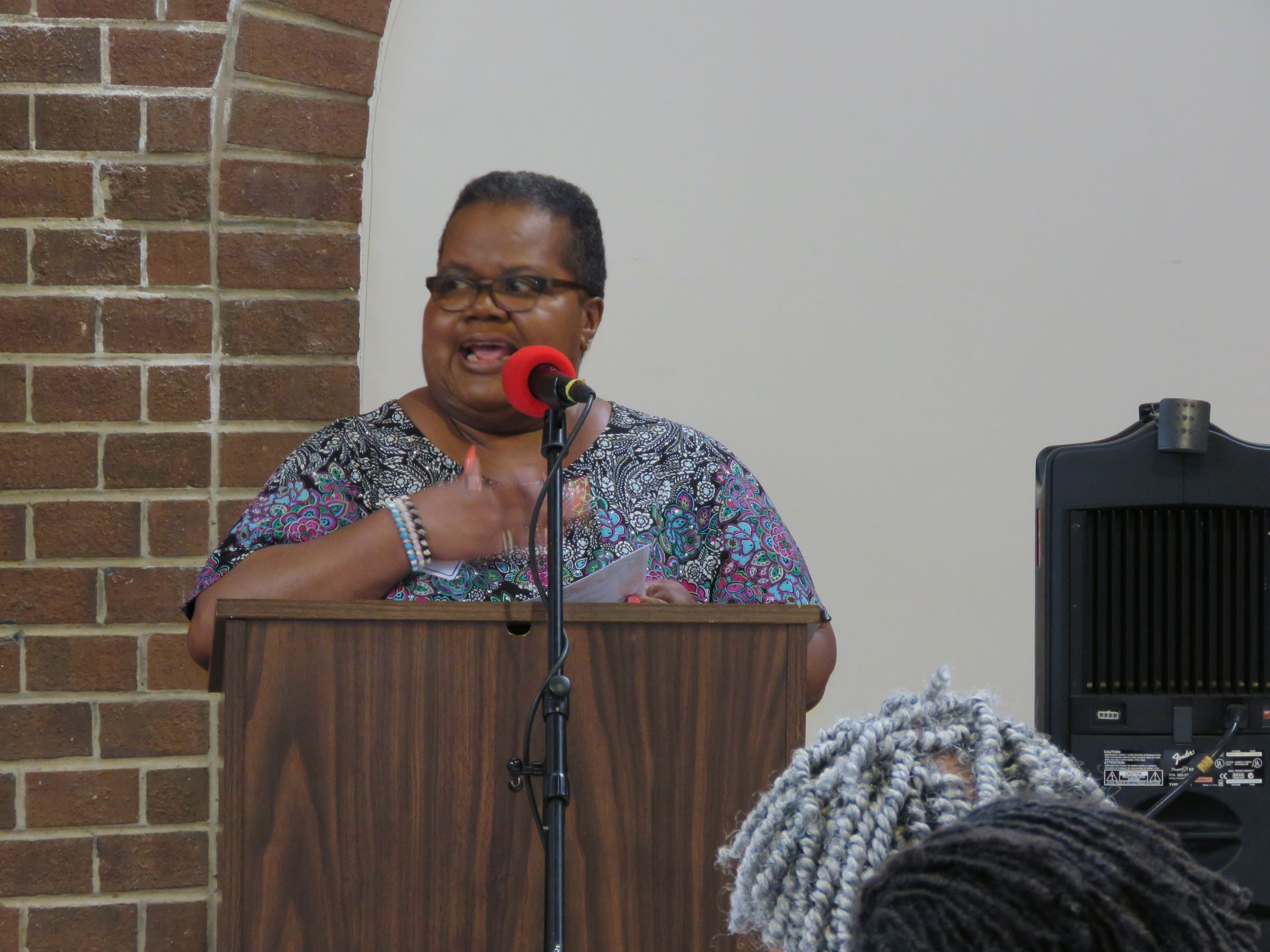 Woman speaking at a lectern, gesturing, microphone in front. Brick wall on the left, speaker to the right.