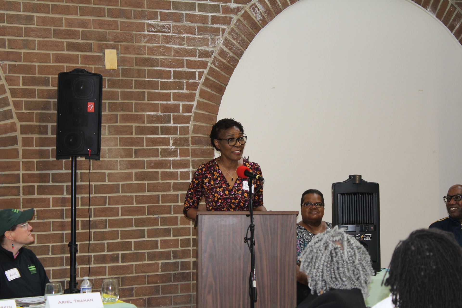 Woman speaking at a podium in a room with a brick wall and seated audience.