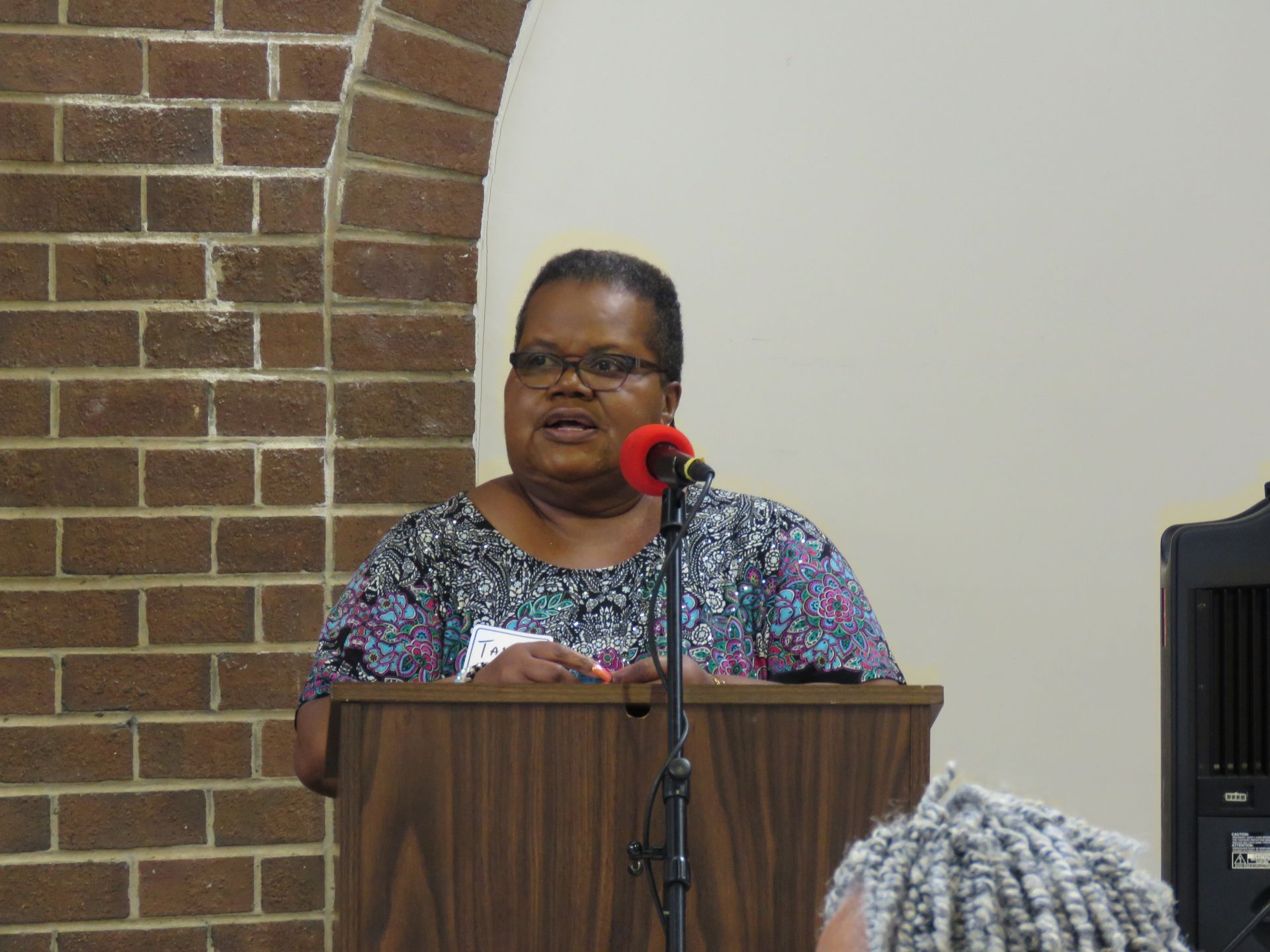 Woman speaking at a podium, brick wall backdrop.