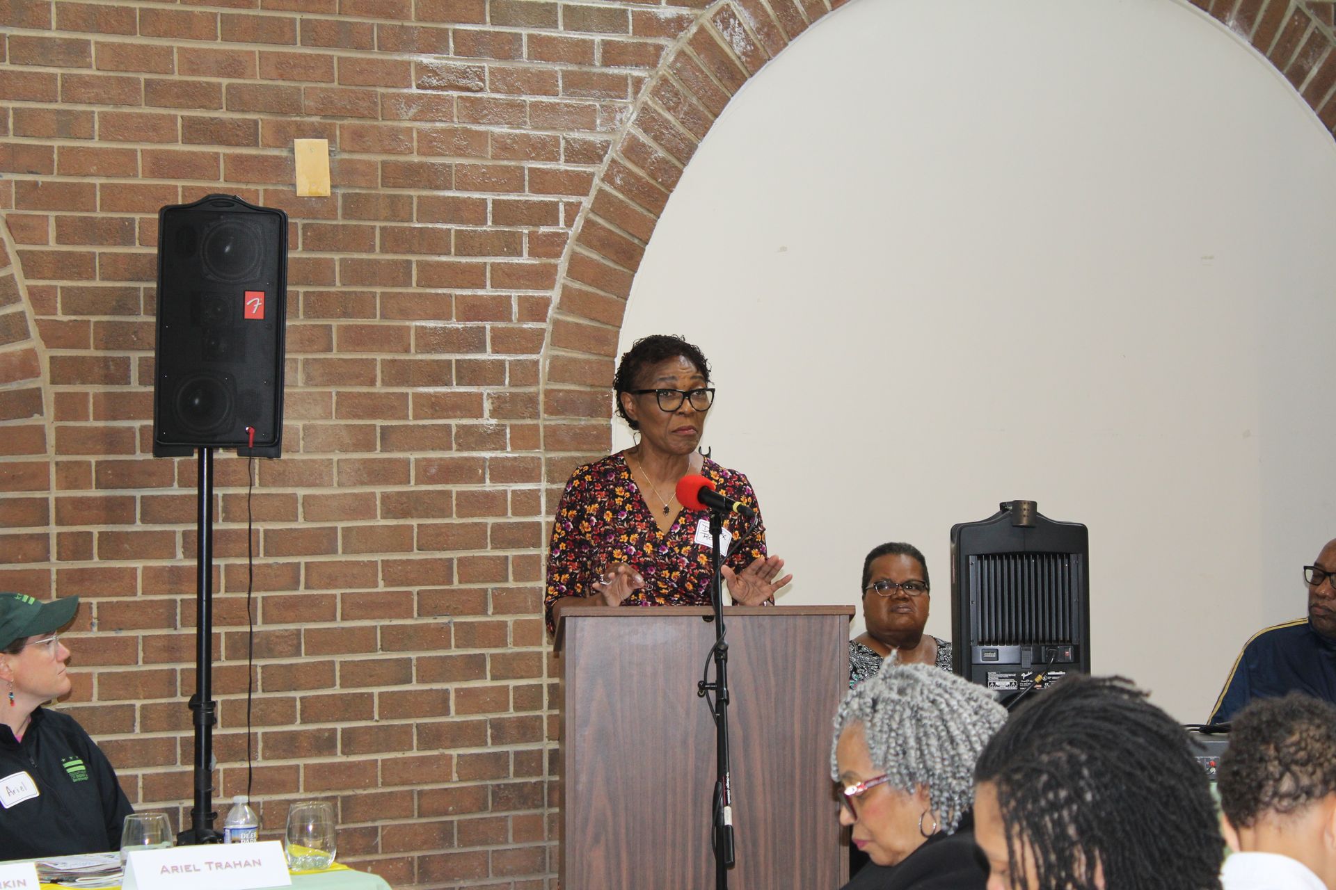 Woman speaking at a podium in a room with a brick wall and arches; an audience sits nearby.