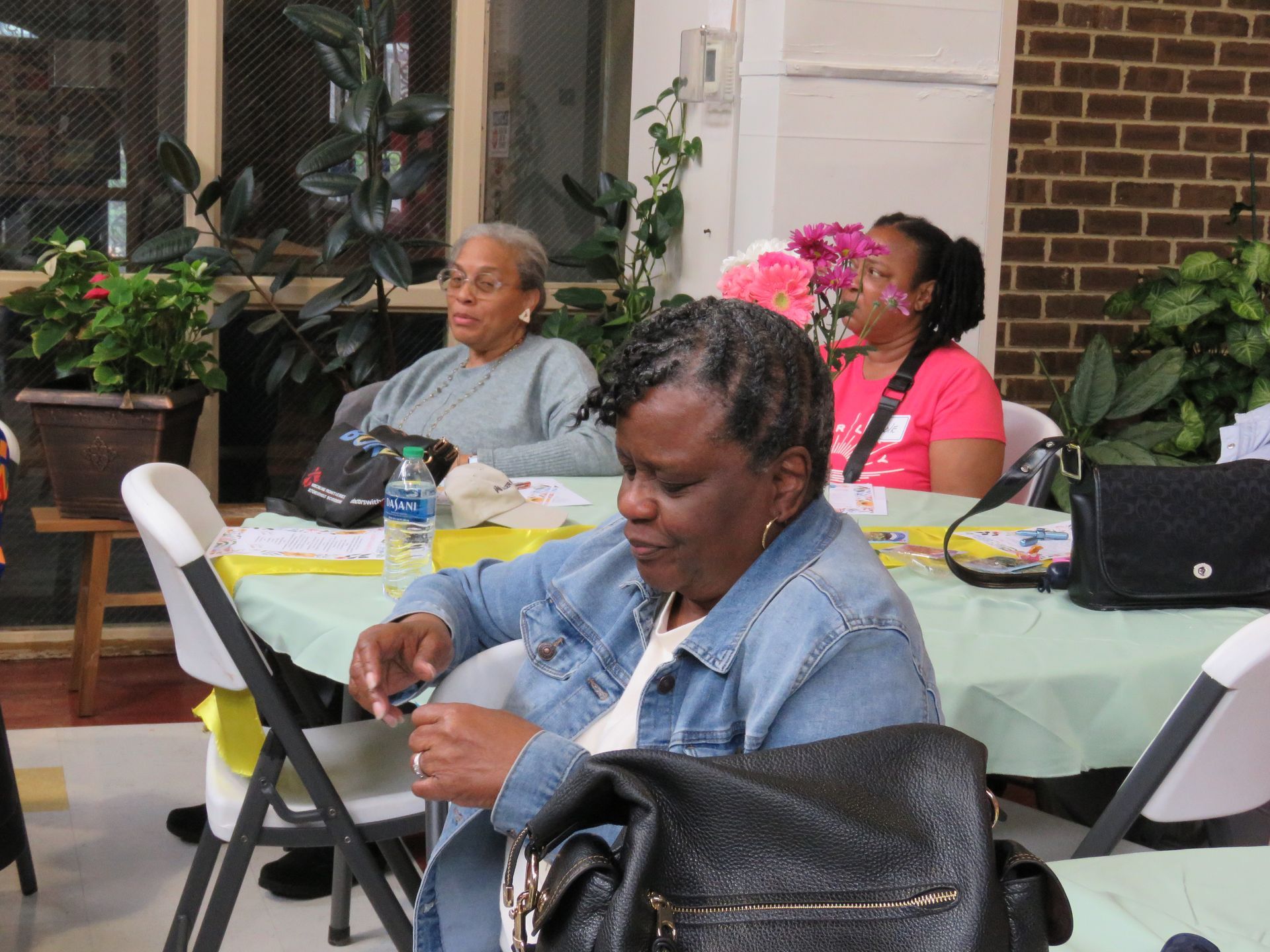 Three women at a table indoors. One in denim jacket, two looking ahead. Plants and brick wall in background.