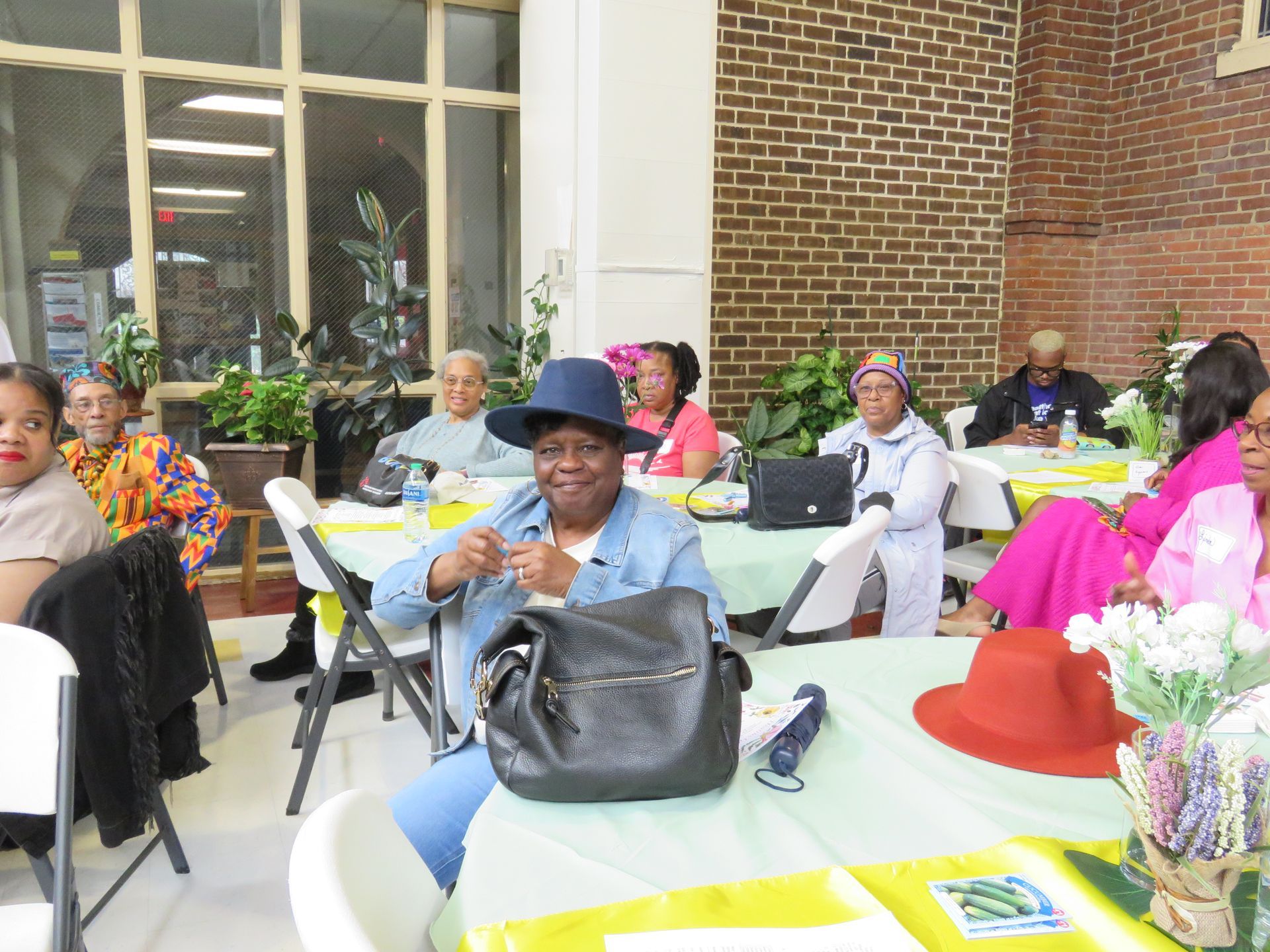 People seated at tables in a room with a brick wall, some smiling, and some are wearing hats.