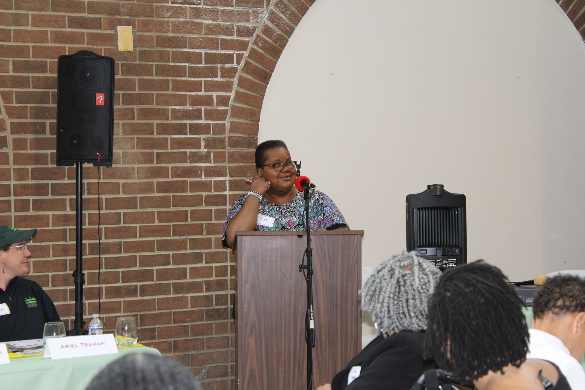 Woman at a podium speaks. Brick wall, speaker, and audience visible.