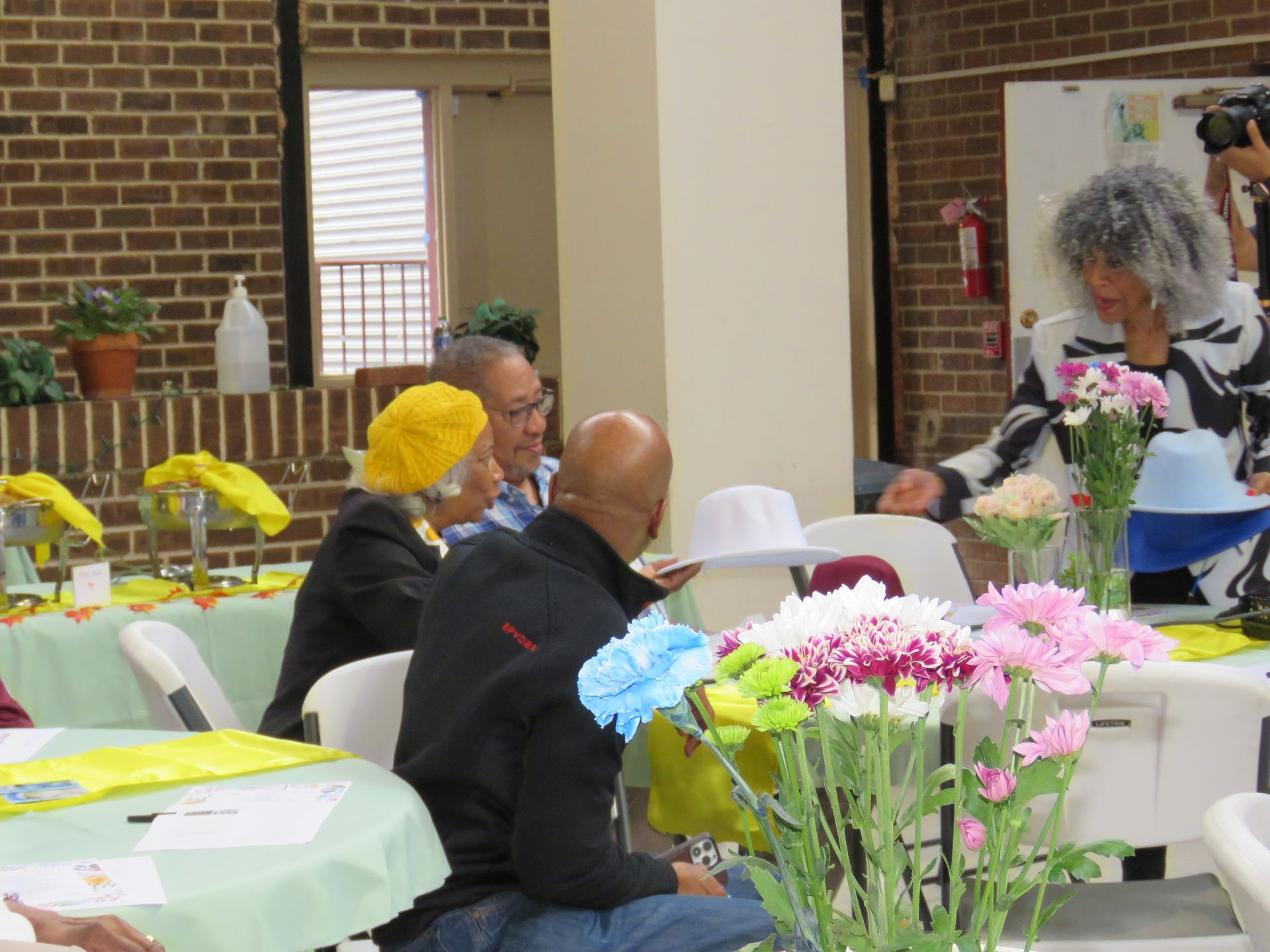 People at a table with flowers, one reaching towards them, brick wall in the background.