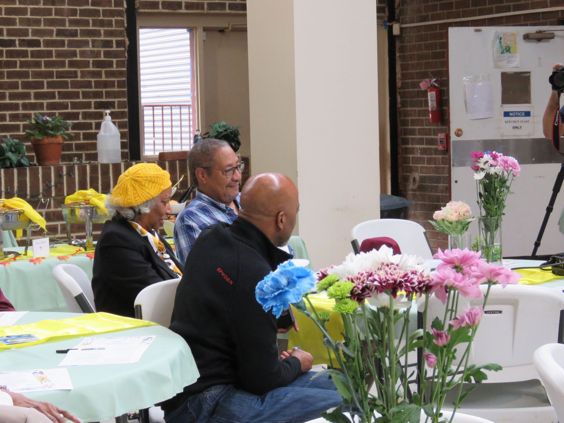 People seated at tables with flowers, indoors. Brick and cream-colored walls. One person wearing a yellow hat.