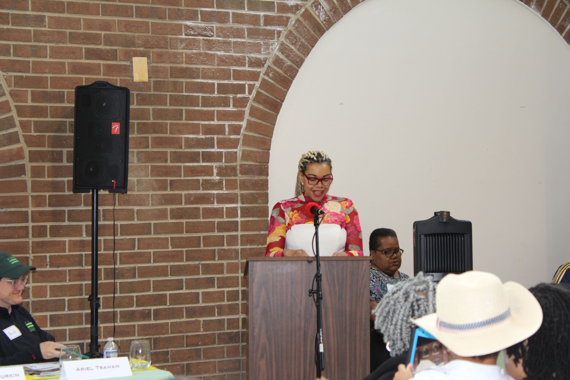 Woman speaking at a podium, brick archway background, people seated at tables, microphone, event setting.