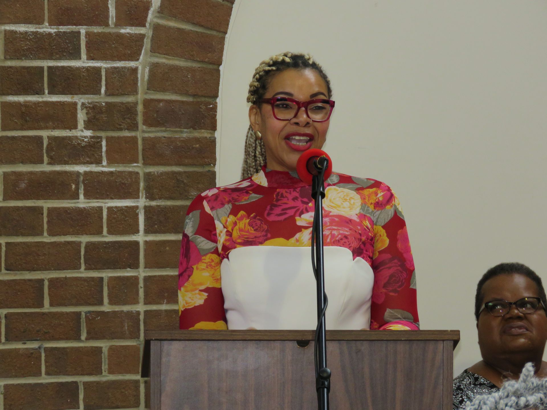 Woman speaking at a podium, wearing glasses, floral top, brick background.