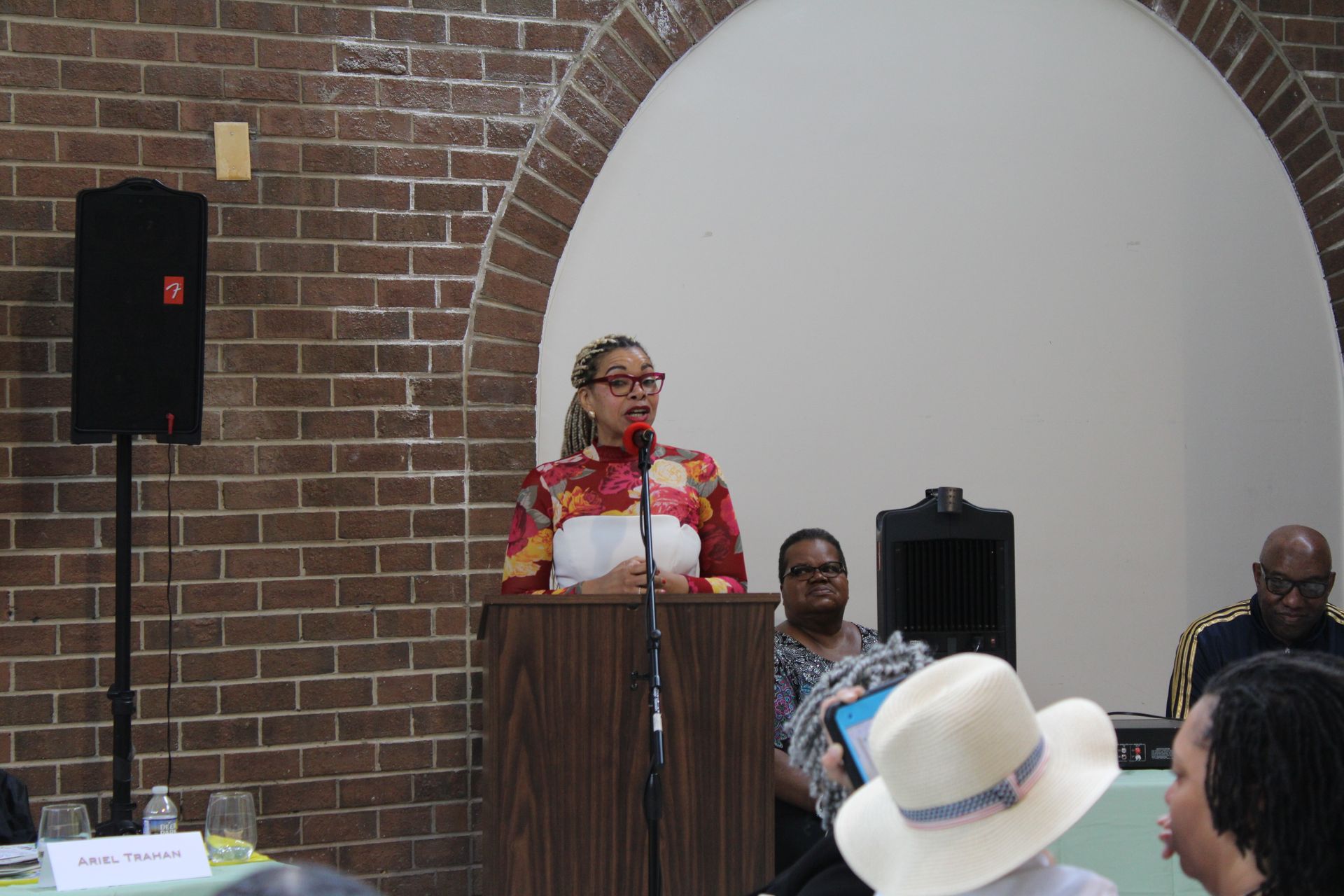 Woman speaking at a podium in a brick-walled room with an arched alcove. Other people are seated.