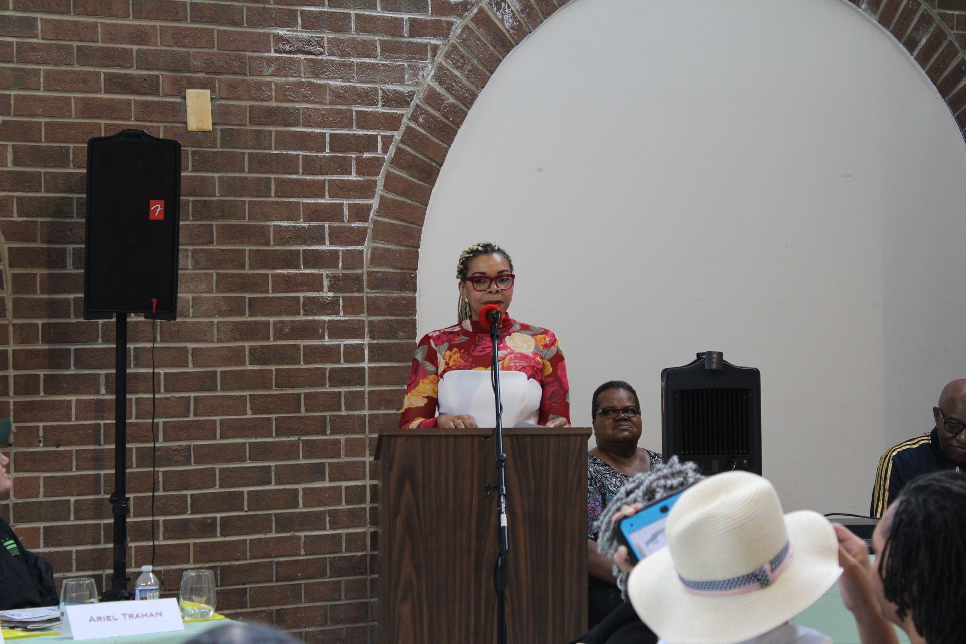 Woman speaking at a podium in a room with a brick arch and audience. She wears a floral patterned shirt.