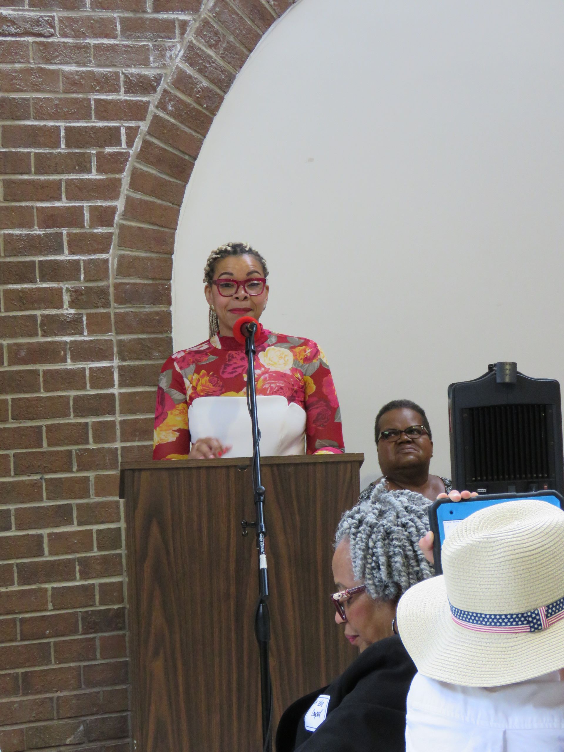 Woman speaking at podium, brick wall backdrop, holding papers, wearing patterned top. Audience present.