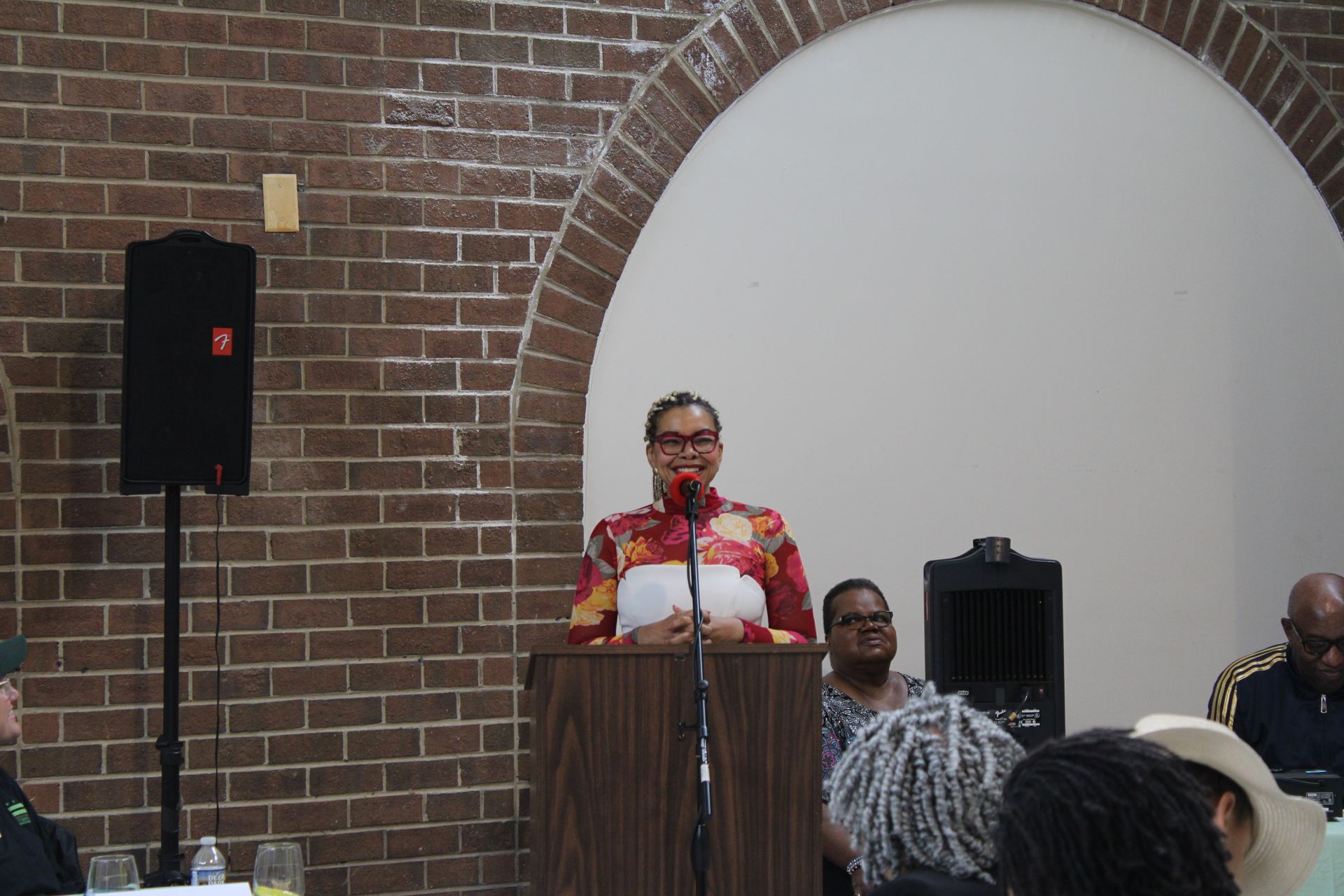 Woman speaking at a podium in a brick-walled room; audience seated.