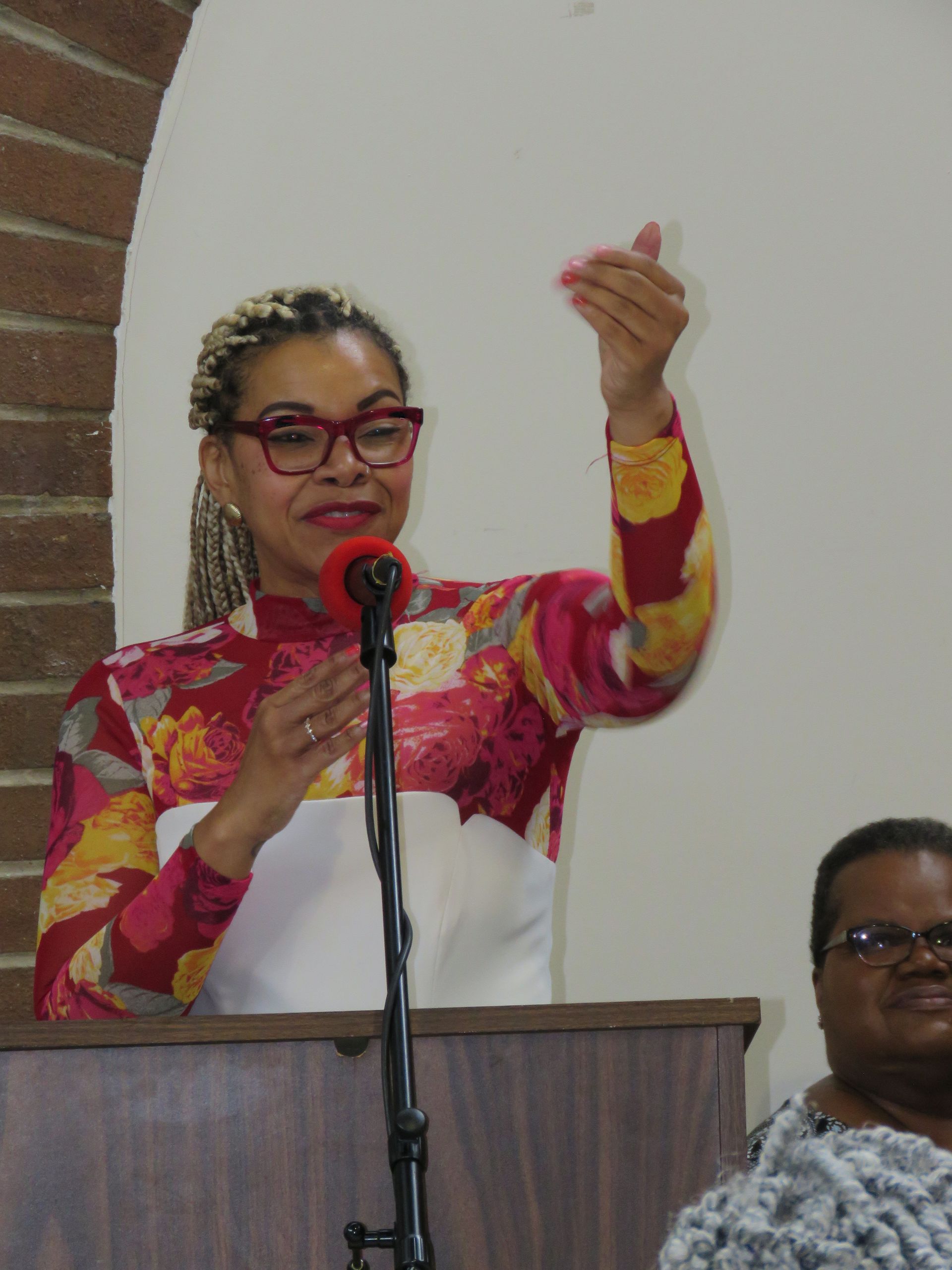 Woman speaking at podium, gesturing with her right hand. Bright floral top, red glasses, and light braids.