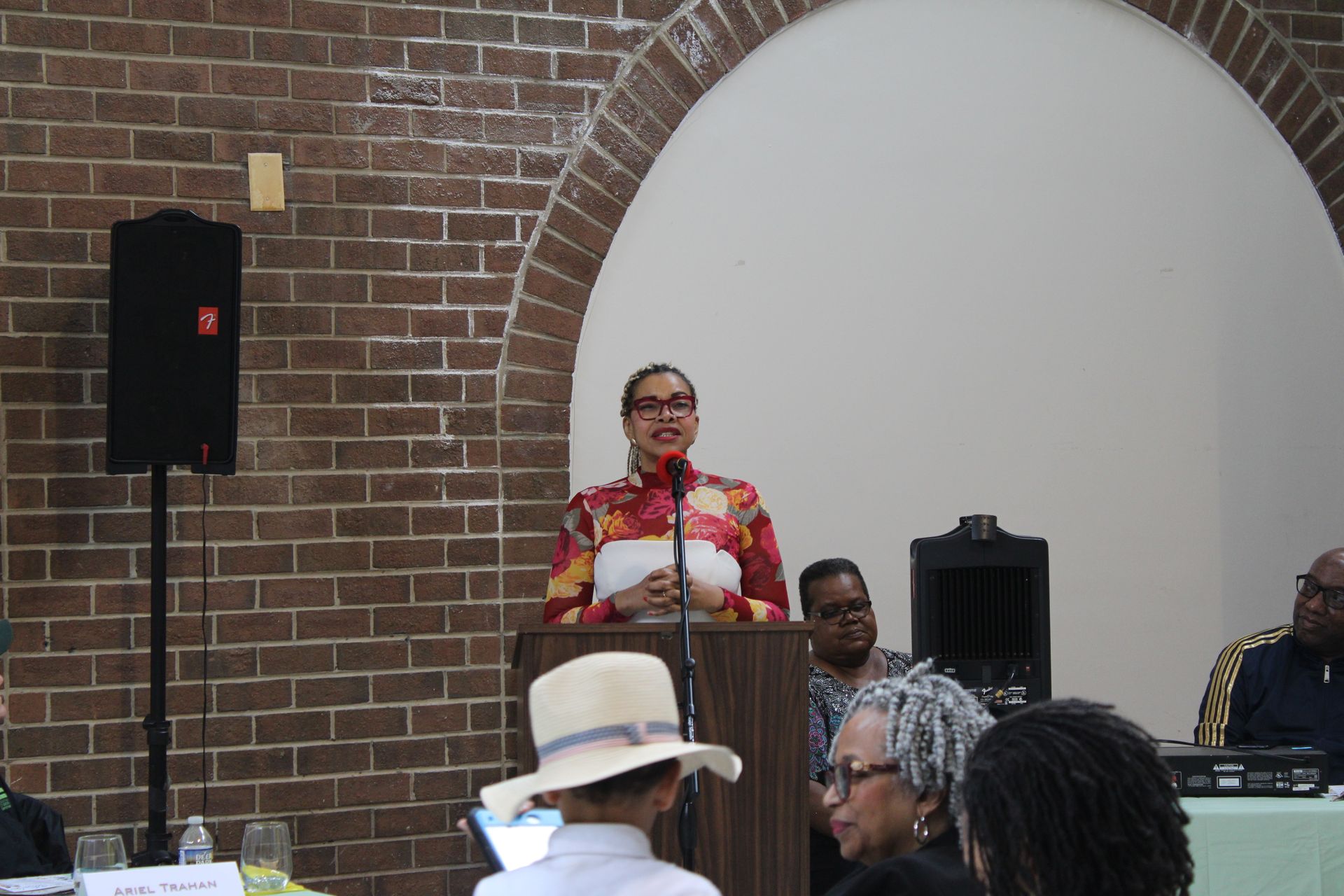 Woman speaking at a podium in a brick-walled room. Attendees listen.