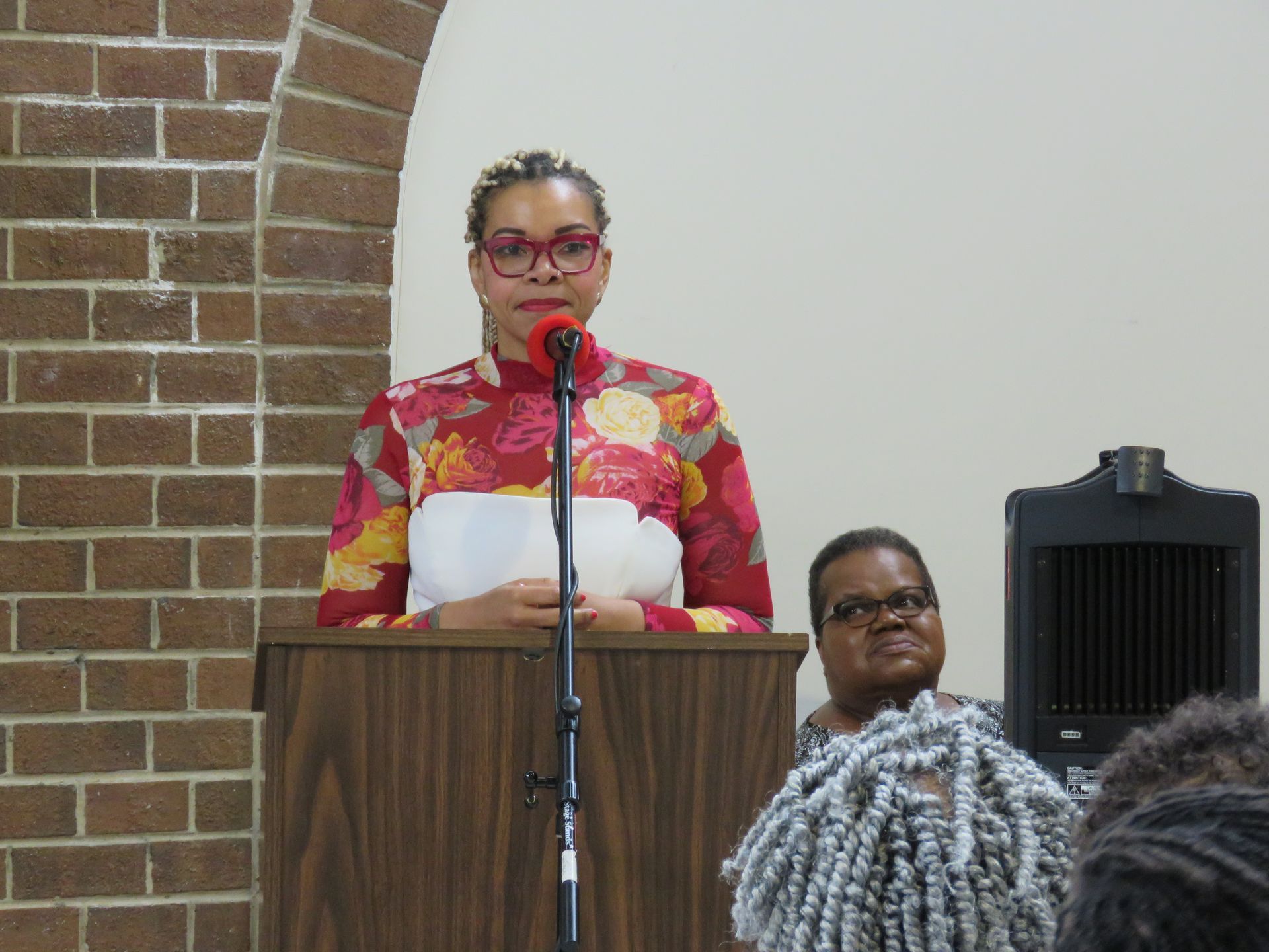 Woman in floral top speaks at podium; another woman sits nearby. Brick wall and beige background.