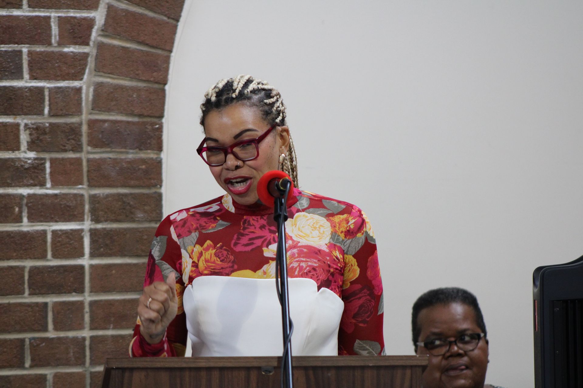 Woman speaking at a lectern with a red floral top, brown glasses, and blonde braids. 