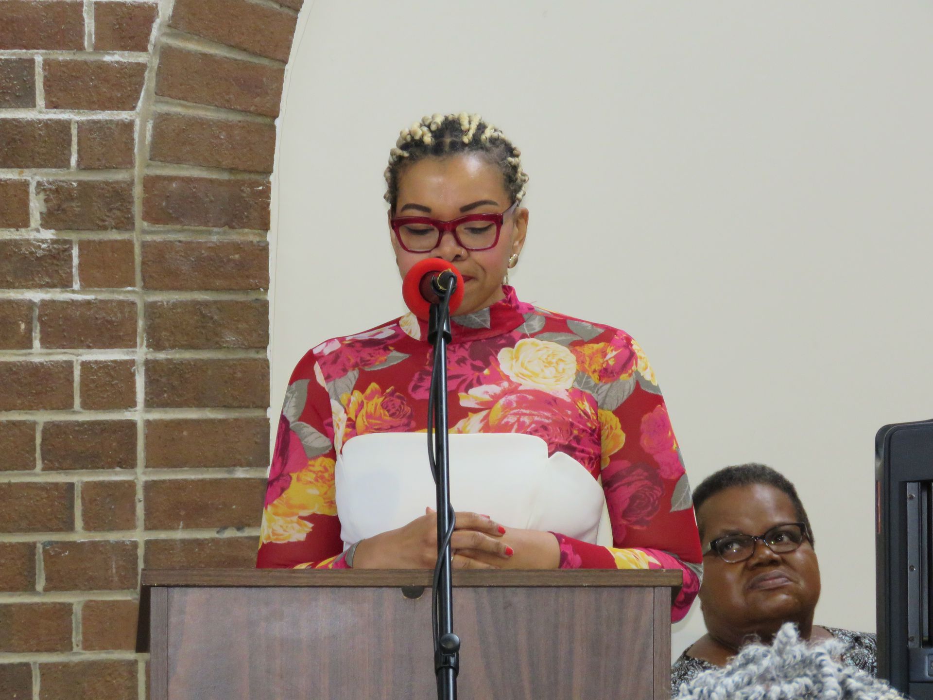Woman speaking at a podium, wearing floral dress, glasses, in a brick-walled setting.