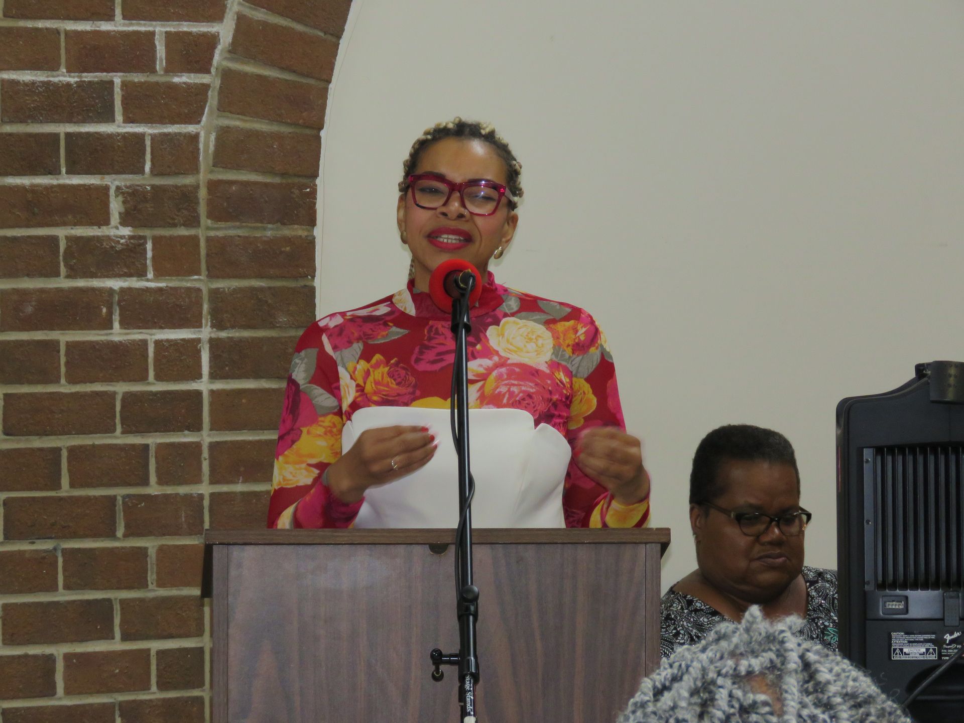 Woman speaking at a podium, wearing floral top and glasses. Brick wall and another woman in background.