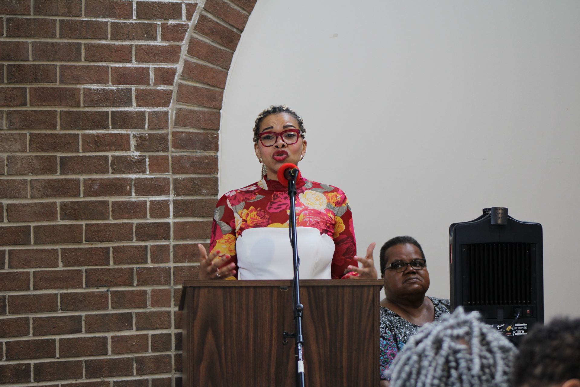 Woman speaking at a podium with floral top, brick wall background.
