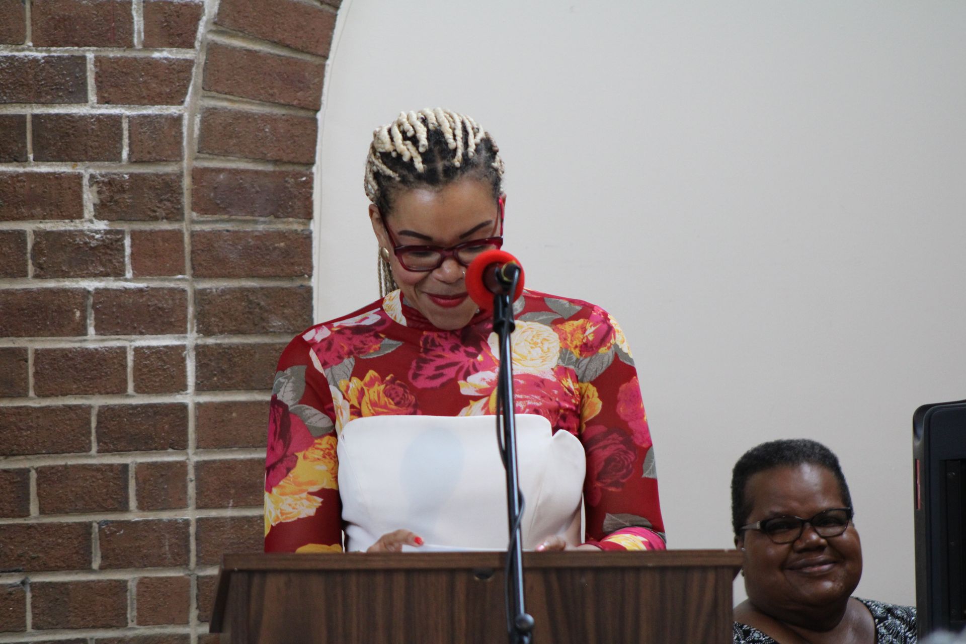 Woman speaking at a podium, wearing glasses and a floral print top, near a brick wall.